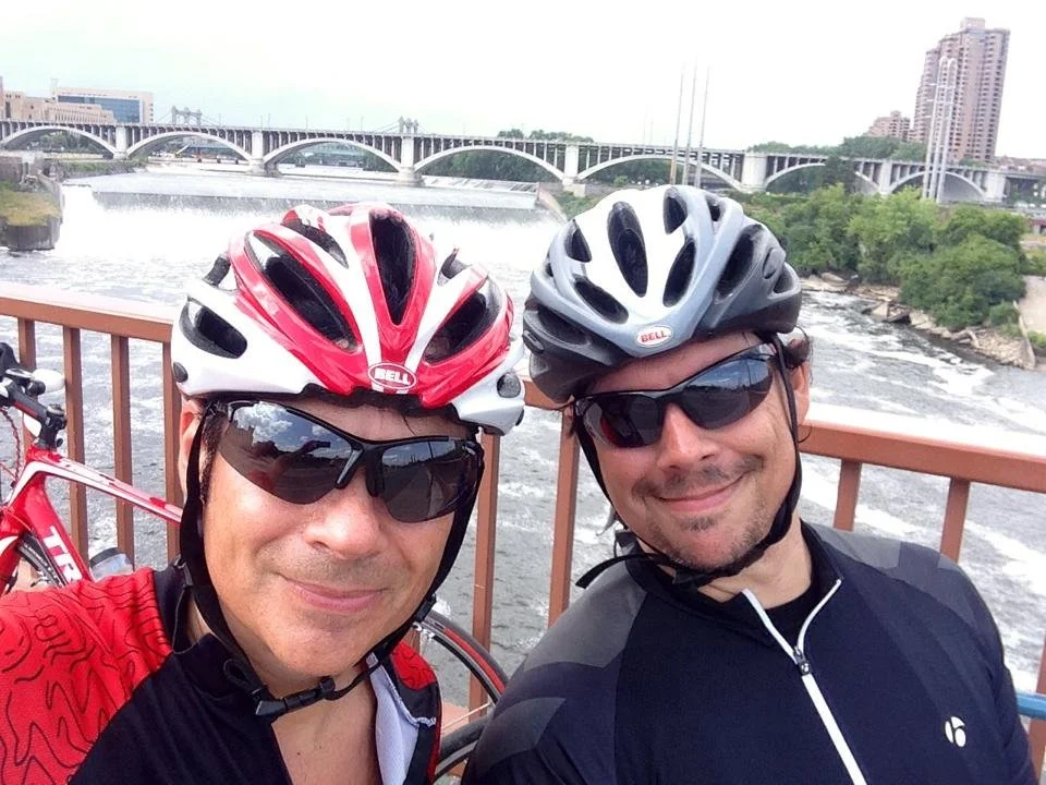 Two men wearing cycling helmets and sunglasses taking a selfie on a bridge with a river and city buildings in the background.