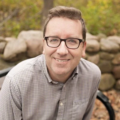 Portrait of a man with glasses smiling outdoors with a rocky background and fall foliage.