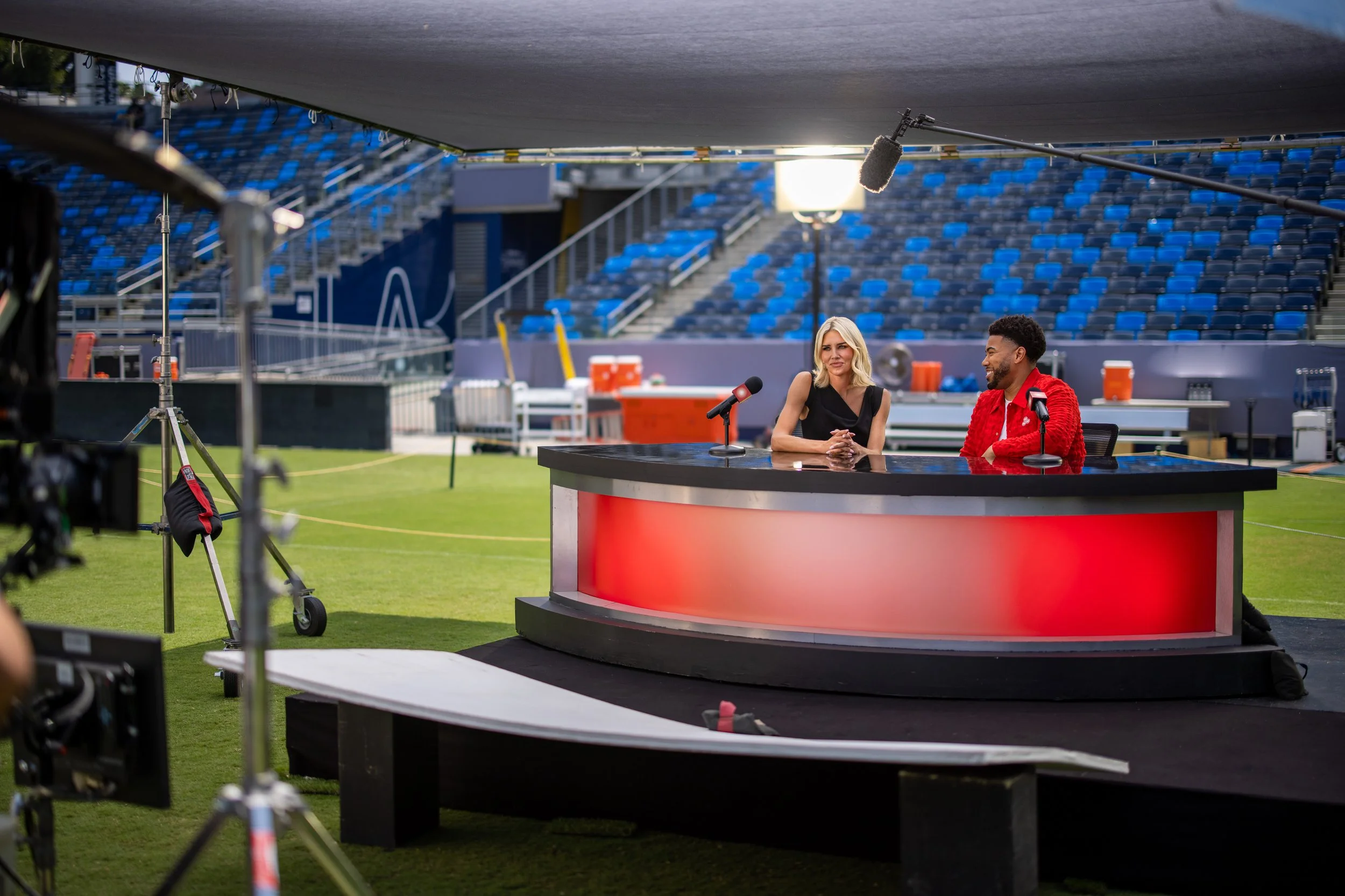 Two hosts sitting at a curved broadcast desk on a sports stadium field, with equipment and cameras nearby, preparing for a live broadcast.