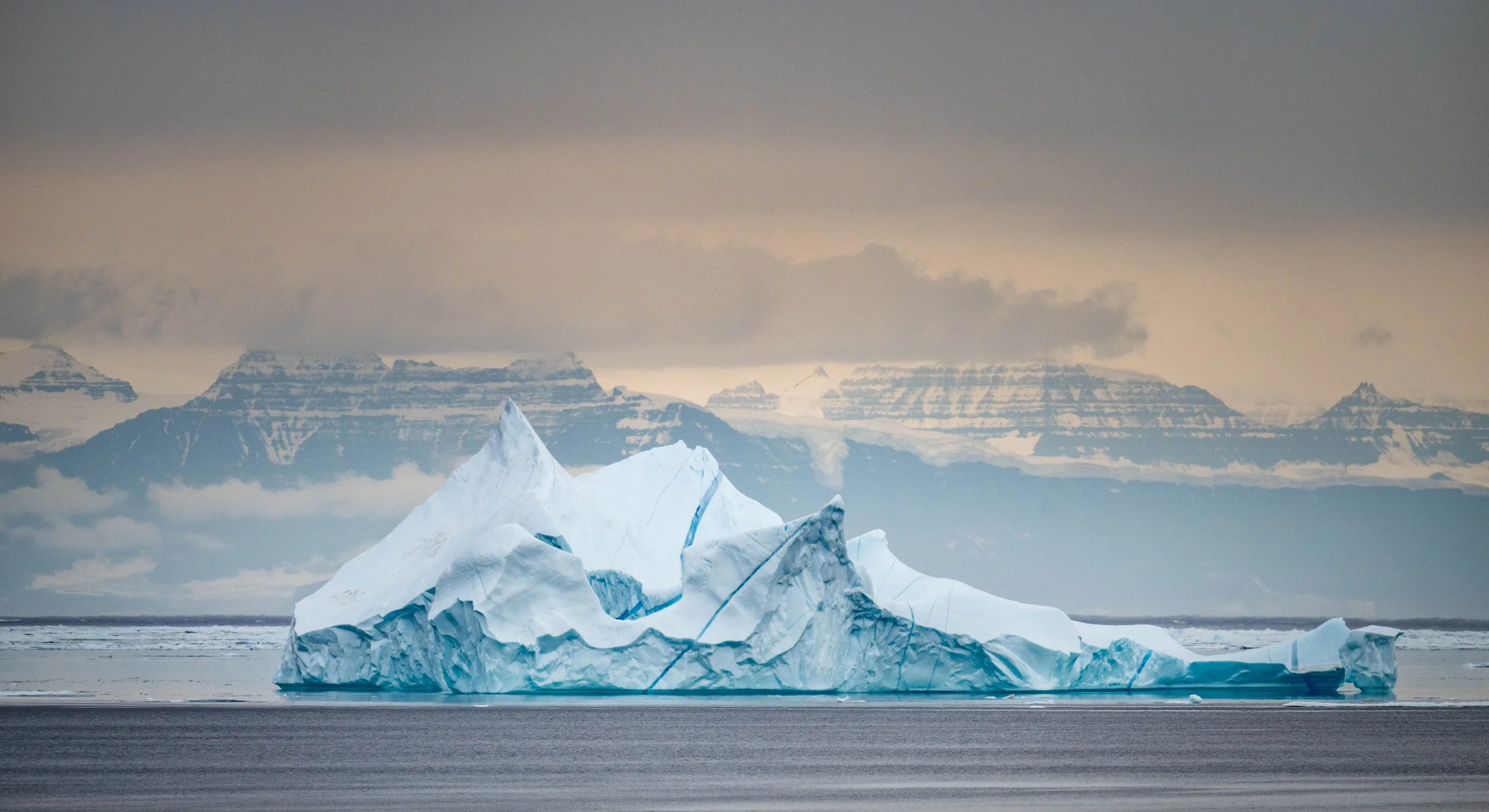 Large iceberg floating in the ocean with snow-covered mountains in the background, under an overcast sky.