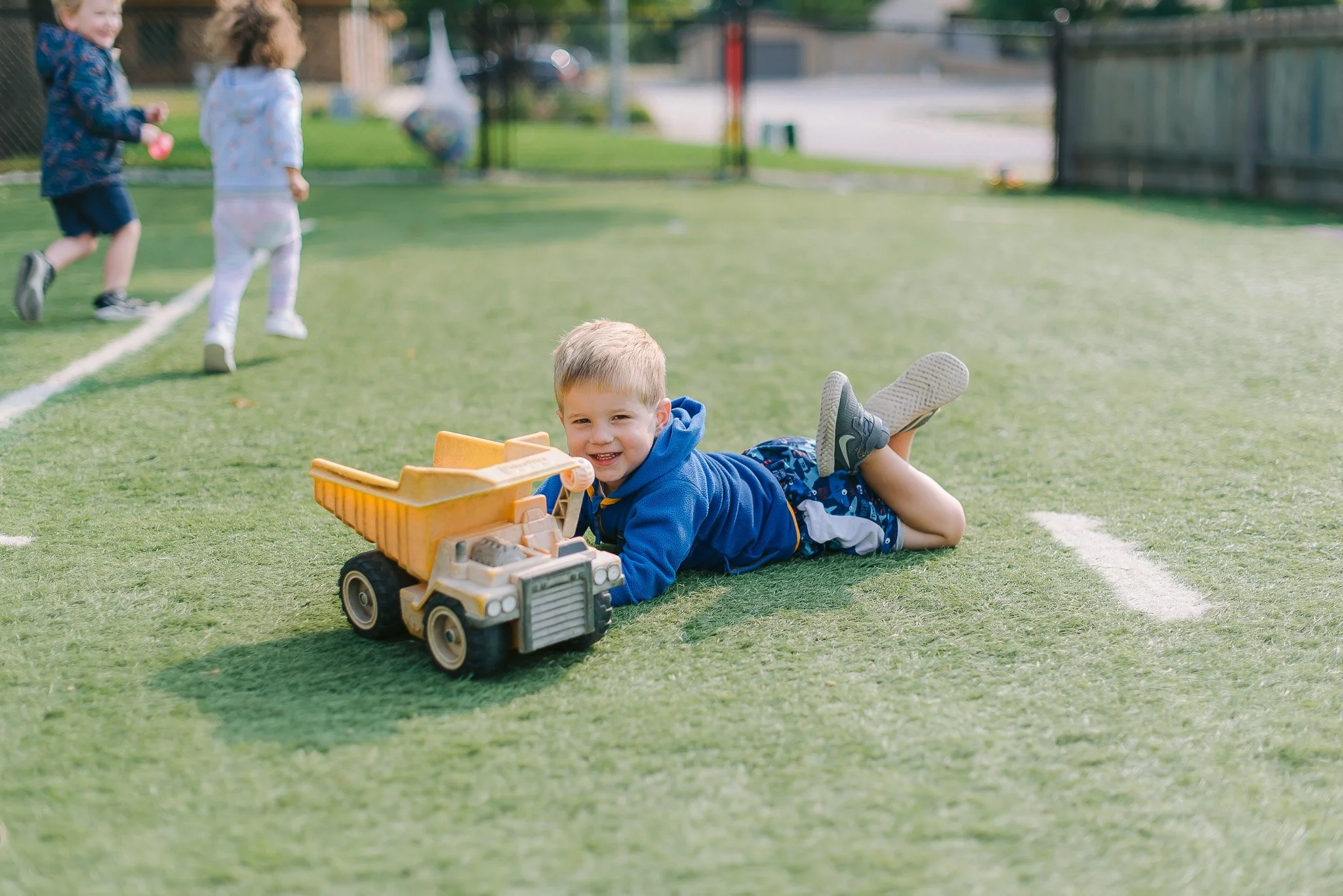 A toddler boy laying down next to a yellow dump truck toy outside