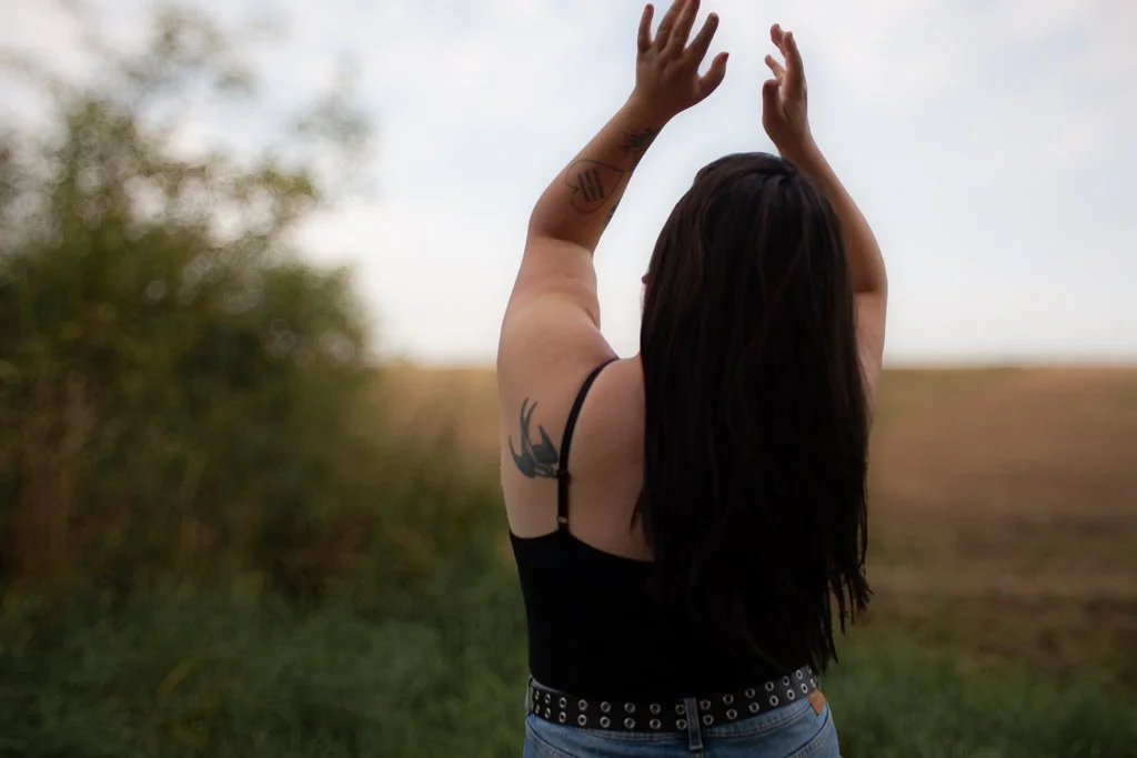 Eine junge Frau mit dunklem Haar steht im Freien, hebt die Arme und blickt in den Himmel.
