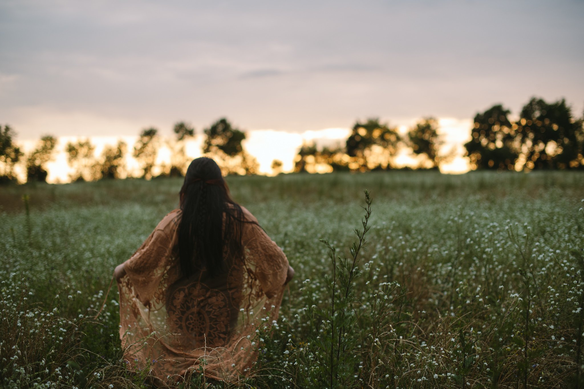 Eine Frau sitzt in einem blühenden Feld bei Sonnenuntergang, mit Blick auf den Himmel und die Bäume im Hintergrund.