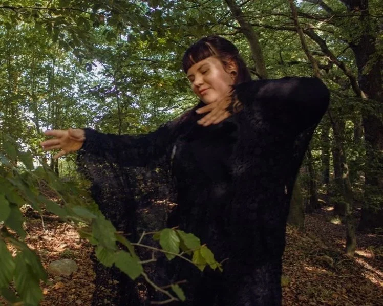 Eine junge Frau in einem schwarzen Kleid steht in einem bewaldeten Gebiet, mit geschlossenen Augen und friedlicher Miene, während sie sich an ihrem Hals berührt.