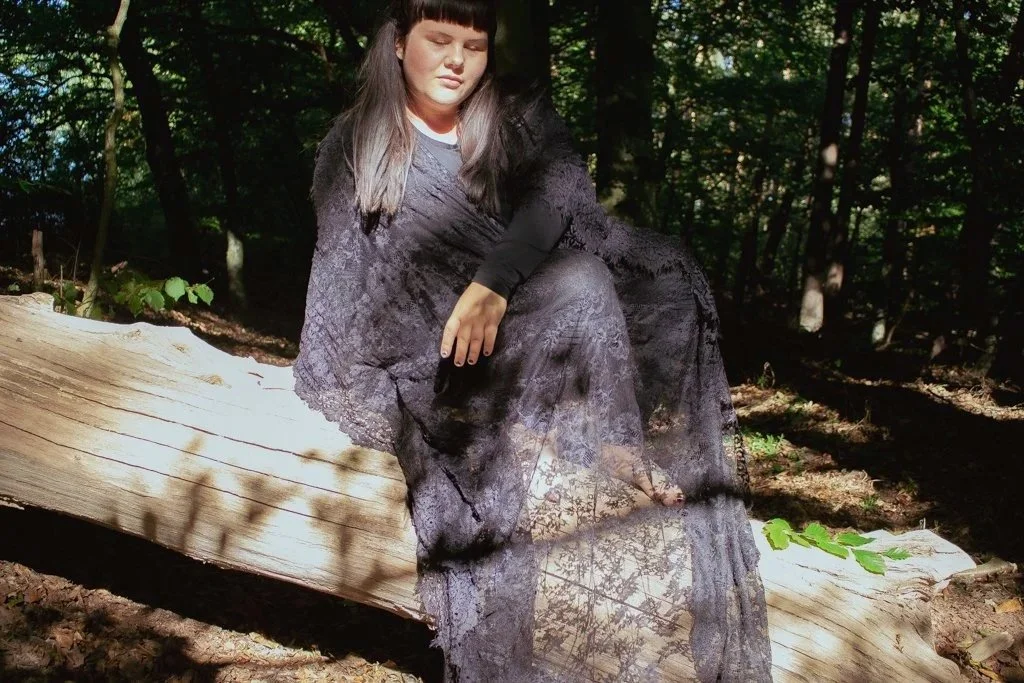 Frau in einem schwarzen Kleid sitzt auf einem Baumstamm im Wald bei Tageslicht.