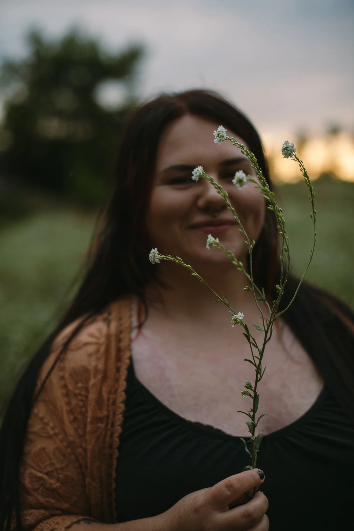 Eine lachende Frau mit langen dunklen Haaren hält eine weiße Blumenstängel vor ihr Gesicht, im Hintergrund ist ein Sonnenuntergang zu sehen.