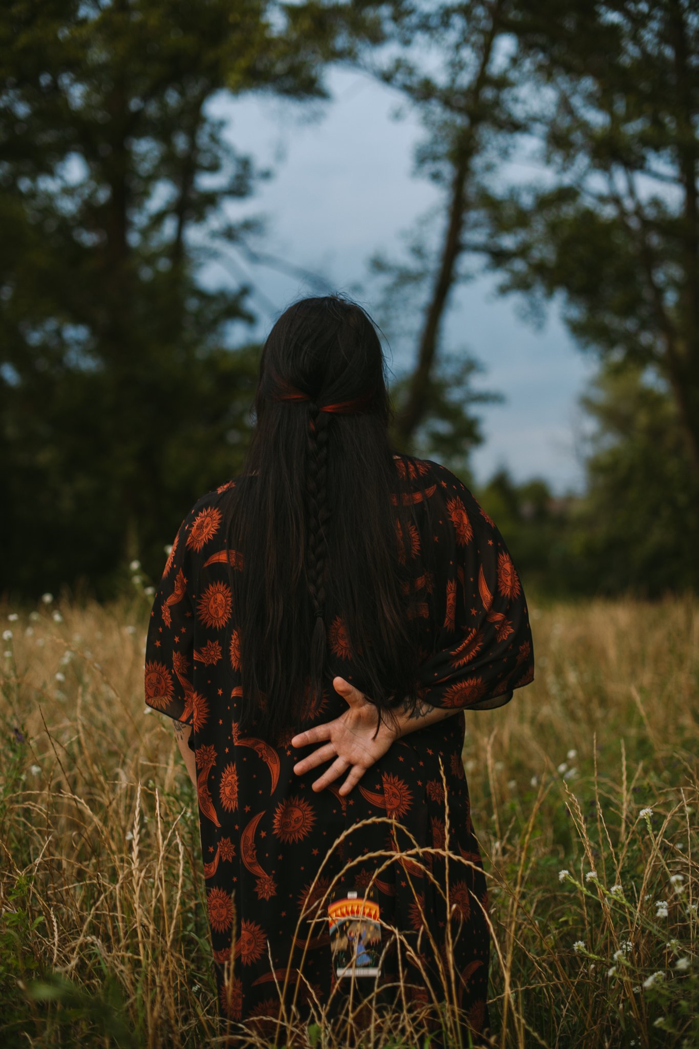 Frau mit langem schwarzen Haar in einem schwarzen Kleid mit oranges Muster steht in einem Feld, hinter Bäume und einen blauen Himmel.