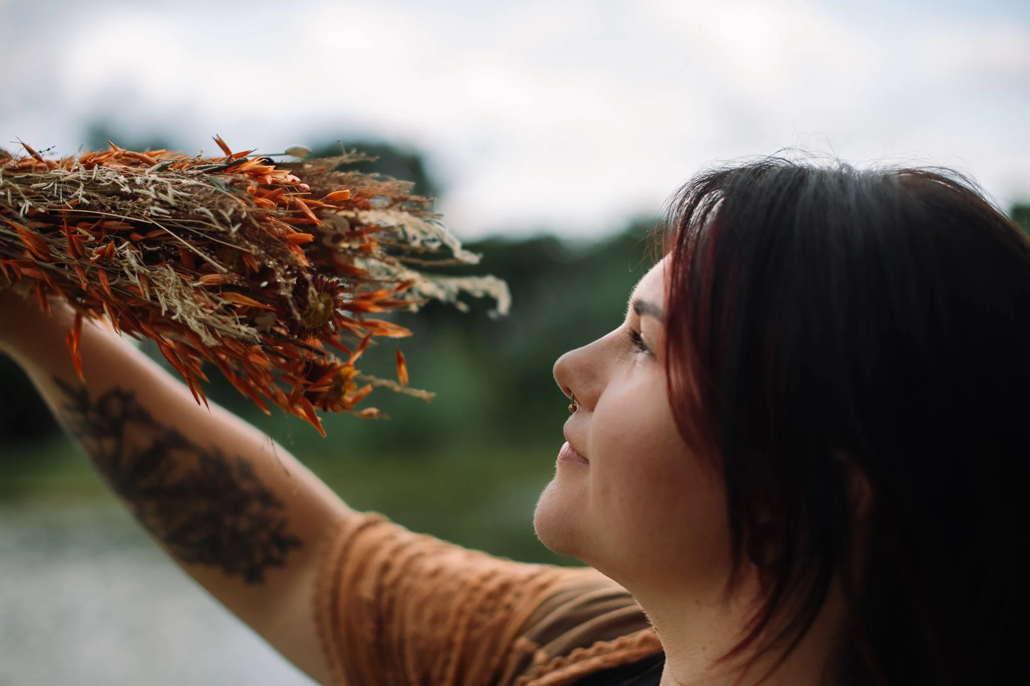 Frau mit dunklem Haar und Tattoos auf dem Arm schaut lächelnd nach oben, hält ein Strauch mit orangem und weißen Blüten gegen den Himmel.