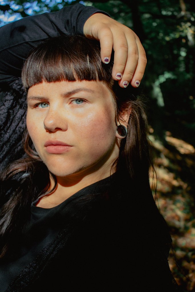 Nahaufnahme eines jungen Mädchens mit blauer Augen, dunklen Haaren mit Pony, der im Wald steht, die Hand auf den Kopf gelegt, dunkler Pullover, Ohrschmuck und dunkle Nägel.