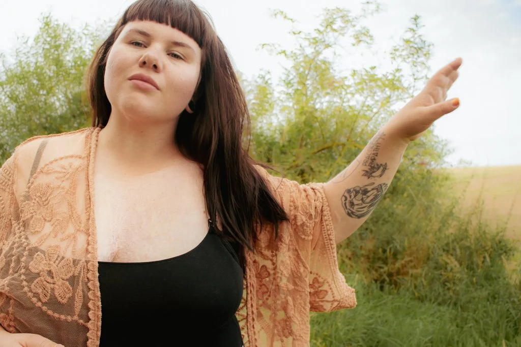 Junge Frau mit dunklen Haaren, tätowiertem Arm, trägt schwarzes Top und eine orangefarbene Spitzenjacke, steht im Freien mit Büschen und Himmel im Hintergrund.