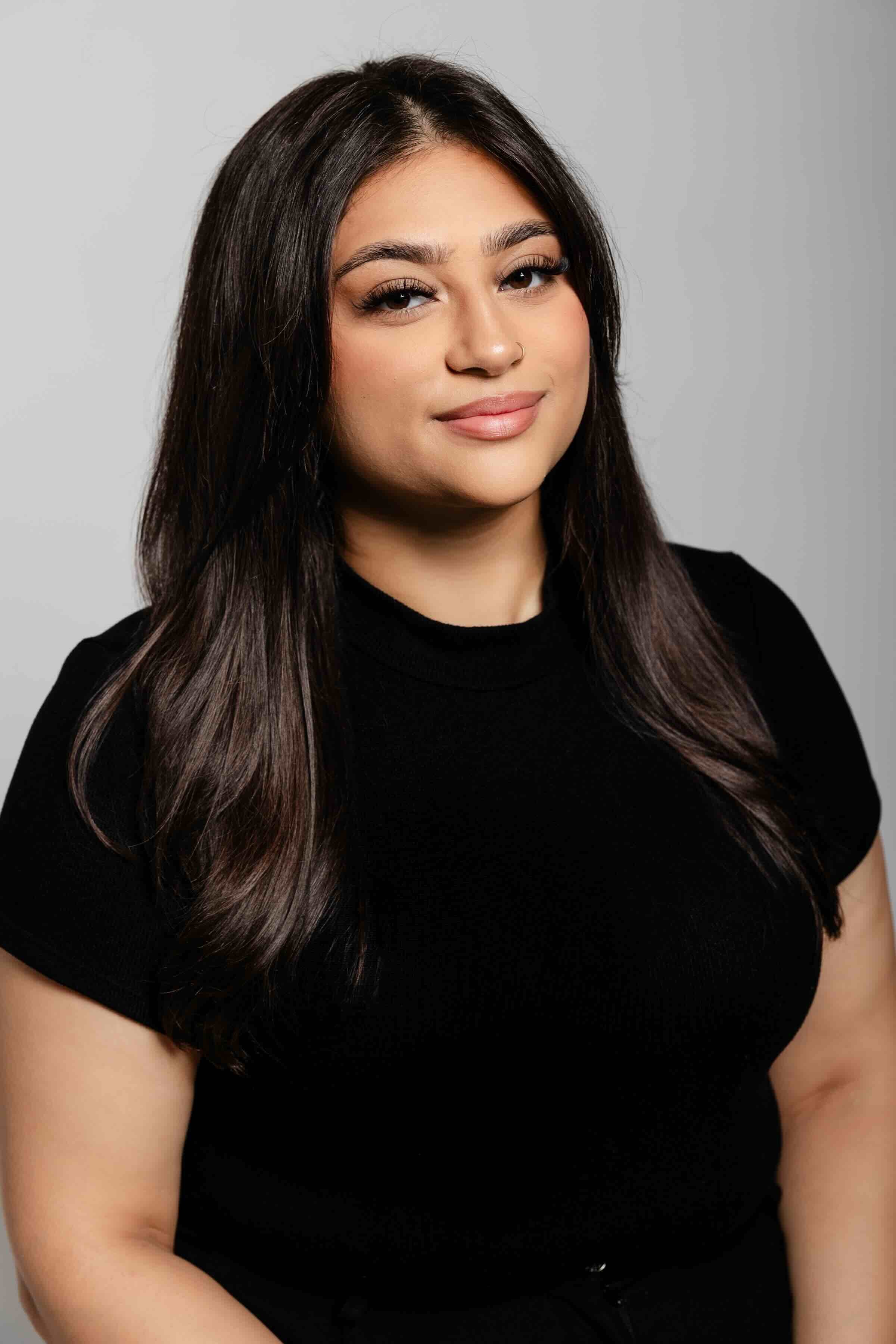 A woman with long dark brown hair and light skin, wearing a black shirt, smiling gently at the camera with a neutral gray background.