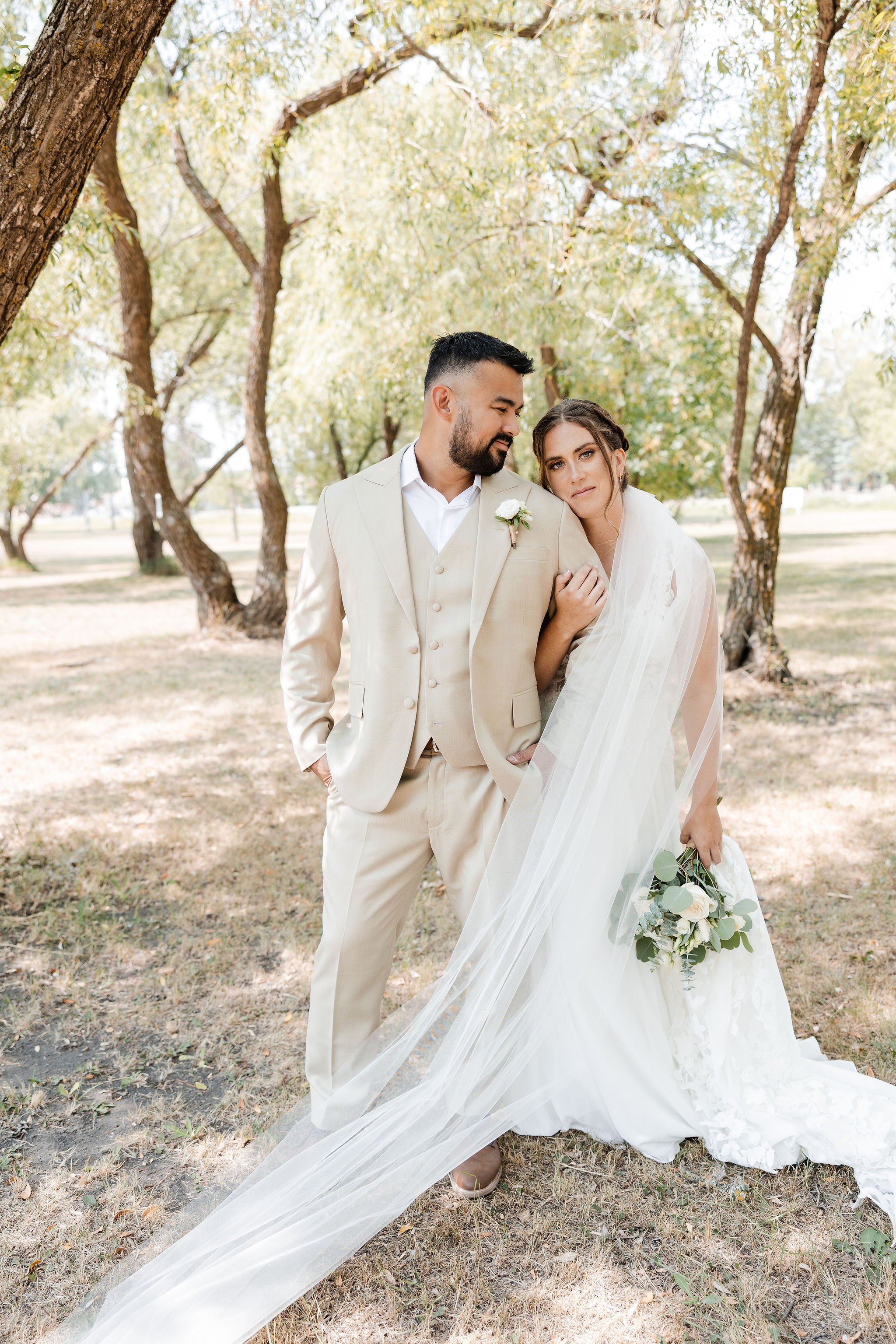 A bride and groom standing together outdoors among trees, with the bride holding a bouquet and the groom with his hand in his pocket, during daytime.