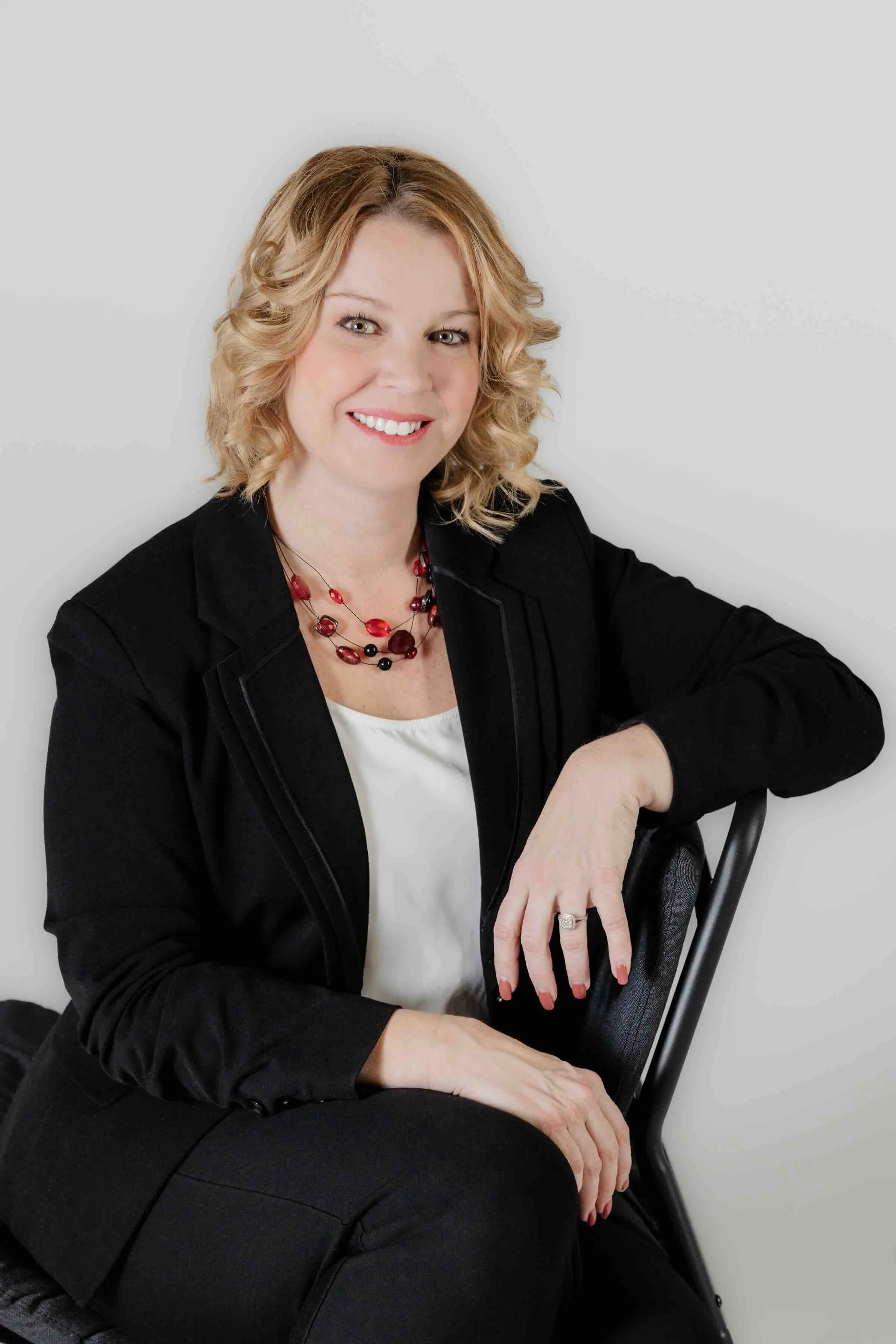 A woman with blonde curly hair smiling, wearing a black blazer and a red beaded necklace, sitting on a black chair against a plain white wall.