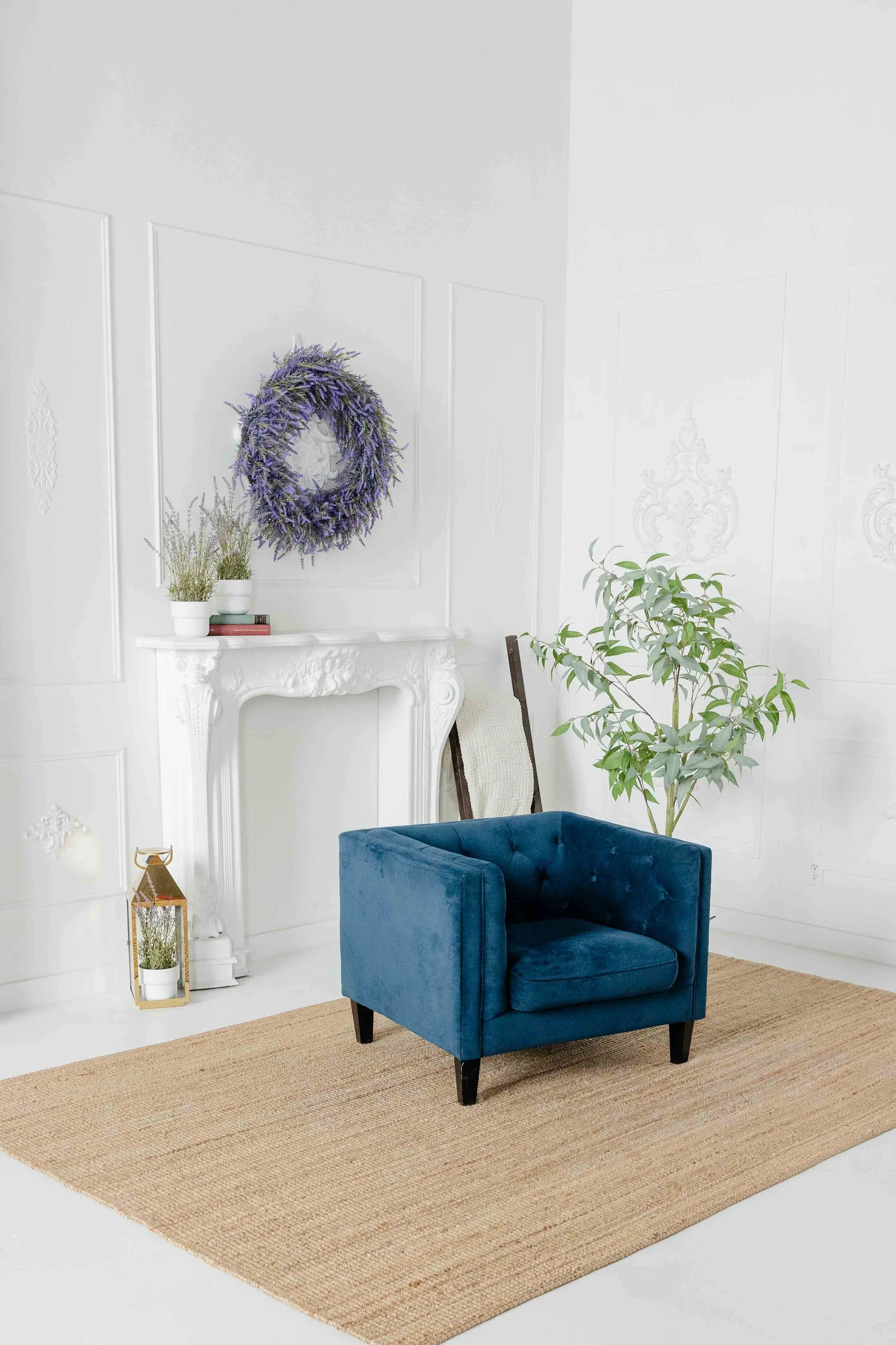 A modern living room with a blue velvet armchair, a large potted plant, a white ornate fireplace mantel, and a lavender wreath hanging above.