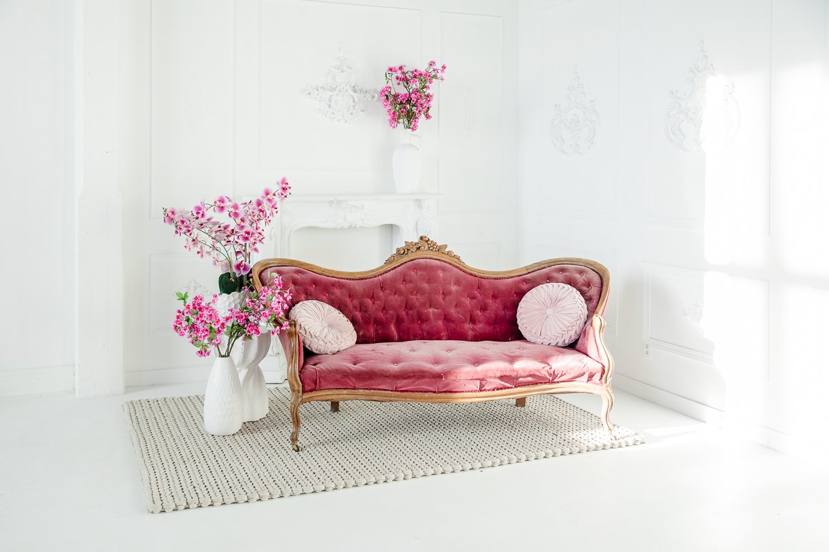 Minimalist living room with white walls and flooring, featuring a blue velvet armchair with a white blanket, a round marble and glass coffee table on a tan flower-shaped rug, a green velvet chair with pink flowering plants in white pots, a fireplace mantel, and a large green plant in a woven basket.