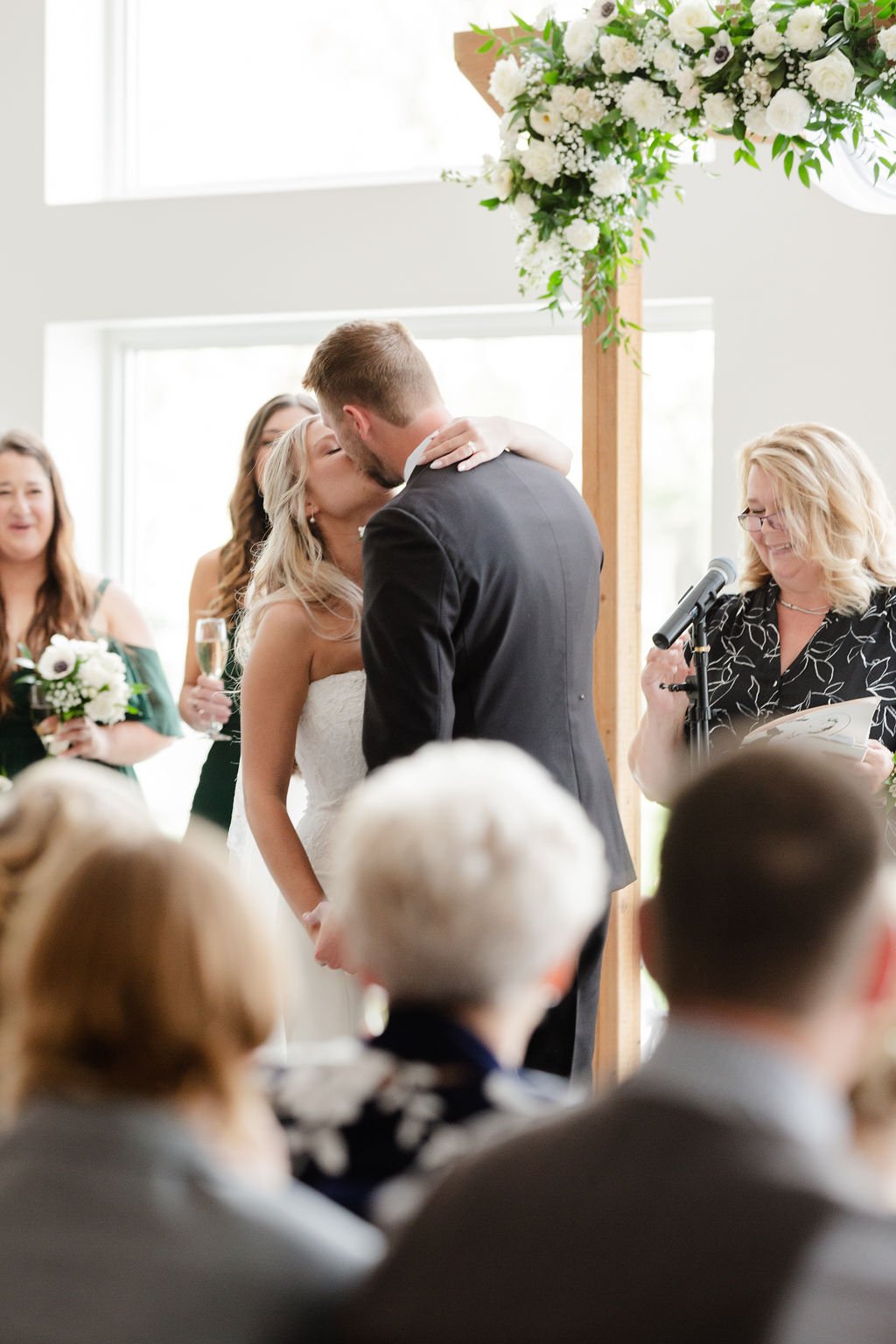 A bride and groom kiss during their wedding ceremony, with officiant and guests present, in a bright venue decorated with white flowers.