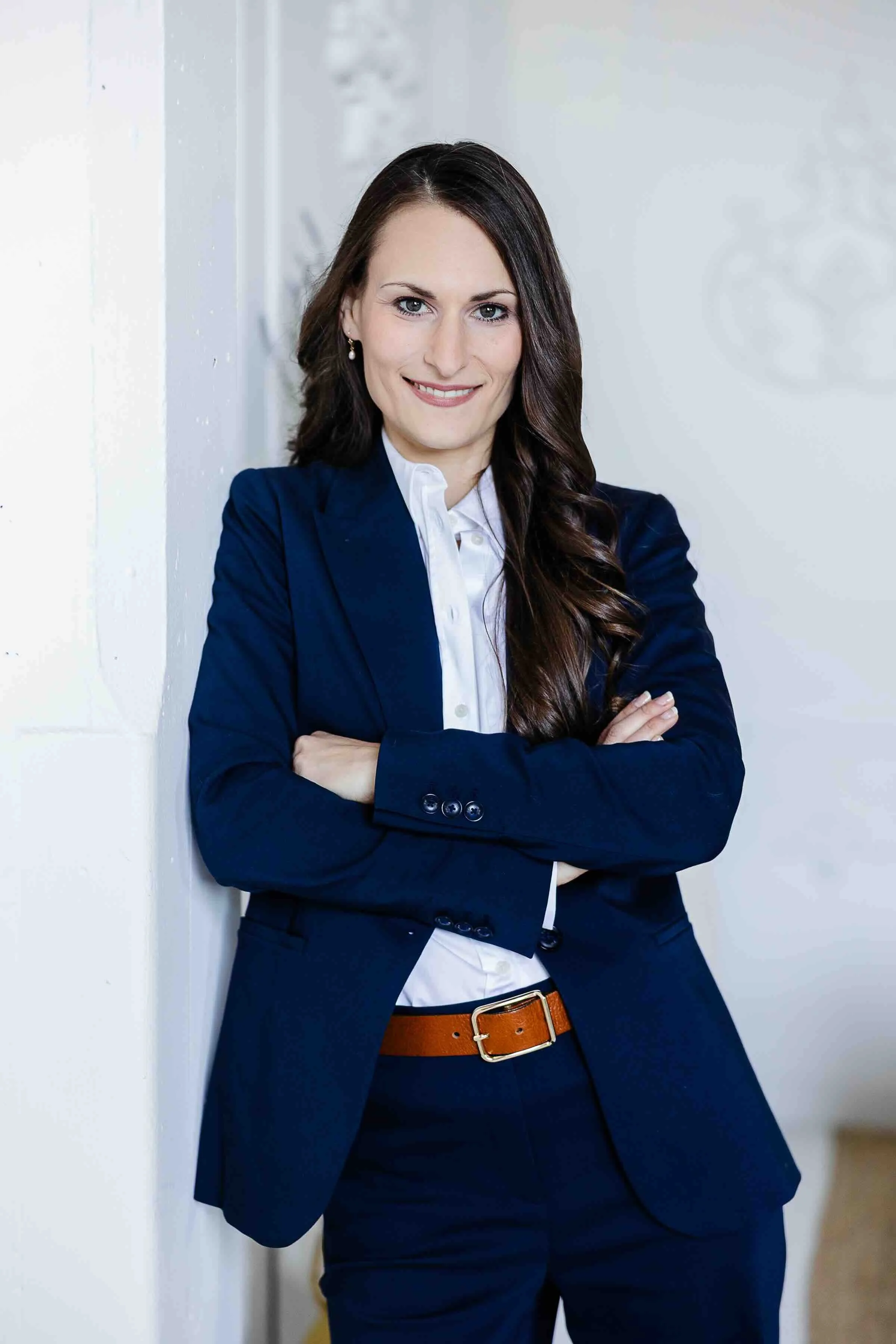 A woman with long dark hair in a blue blazer and white shirt, smiling with arms crossed, standing indoors against a white wall.