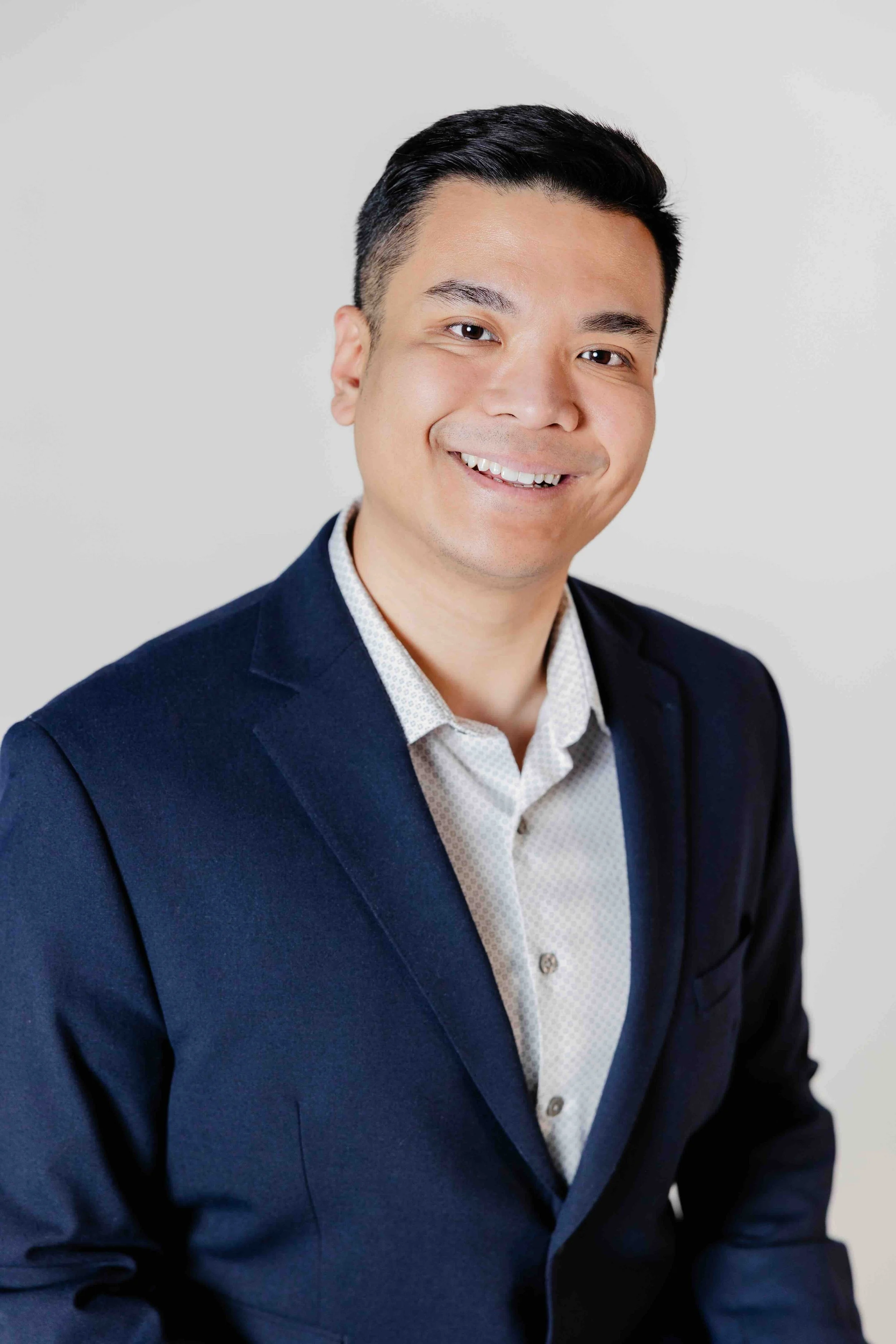 A smiling man in a navy blazer and light-colored shirt poses against a plain light background.