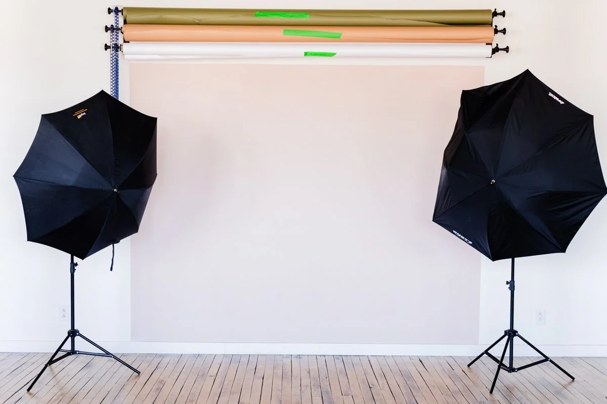 Photography studio setup with two black umbrella lights on stands, a blank backdrop, and various colored rolls of paper or fabric on the ceiling