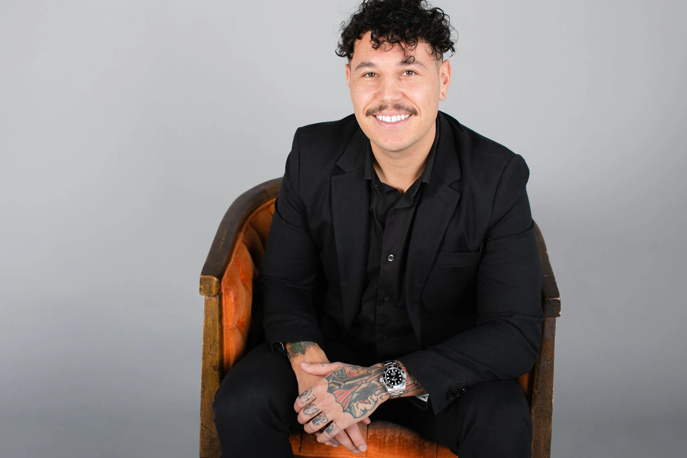 A young man with curly dark hair and a mustache, wearing a black suit and shirt, smiling and sitting on an orange velvet armchair against a plain gray background. He has tattoos on his left hand and wrist, and is wearing a silver watch.