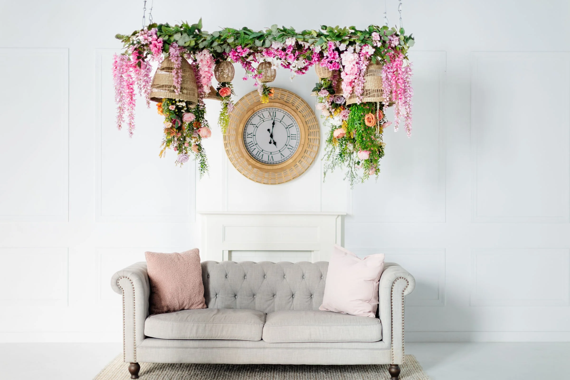 A woman standing in a bright living room holding a laptop, with a large clock on the wall behind her and a potted plant to her left.