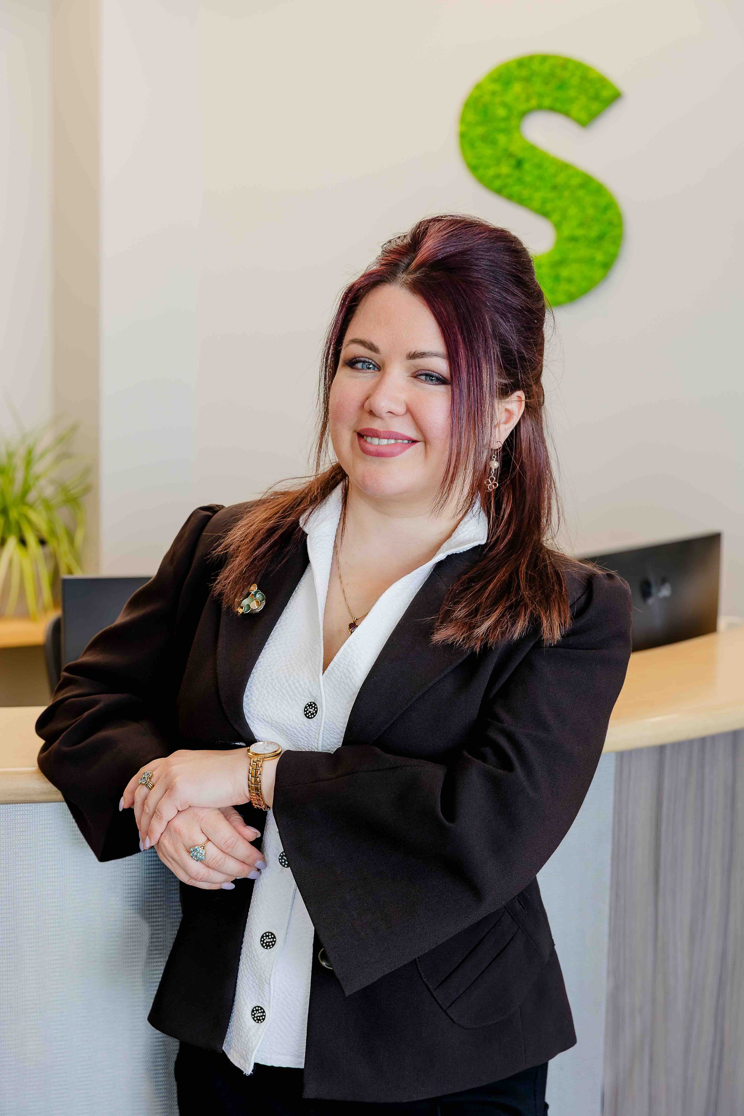 A woman in a black blazer and white blouse standing at a reception desk with a green letter S on the wall behind her.