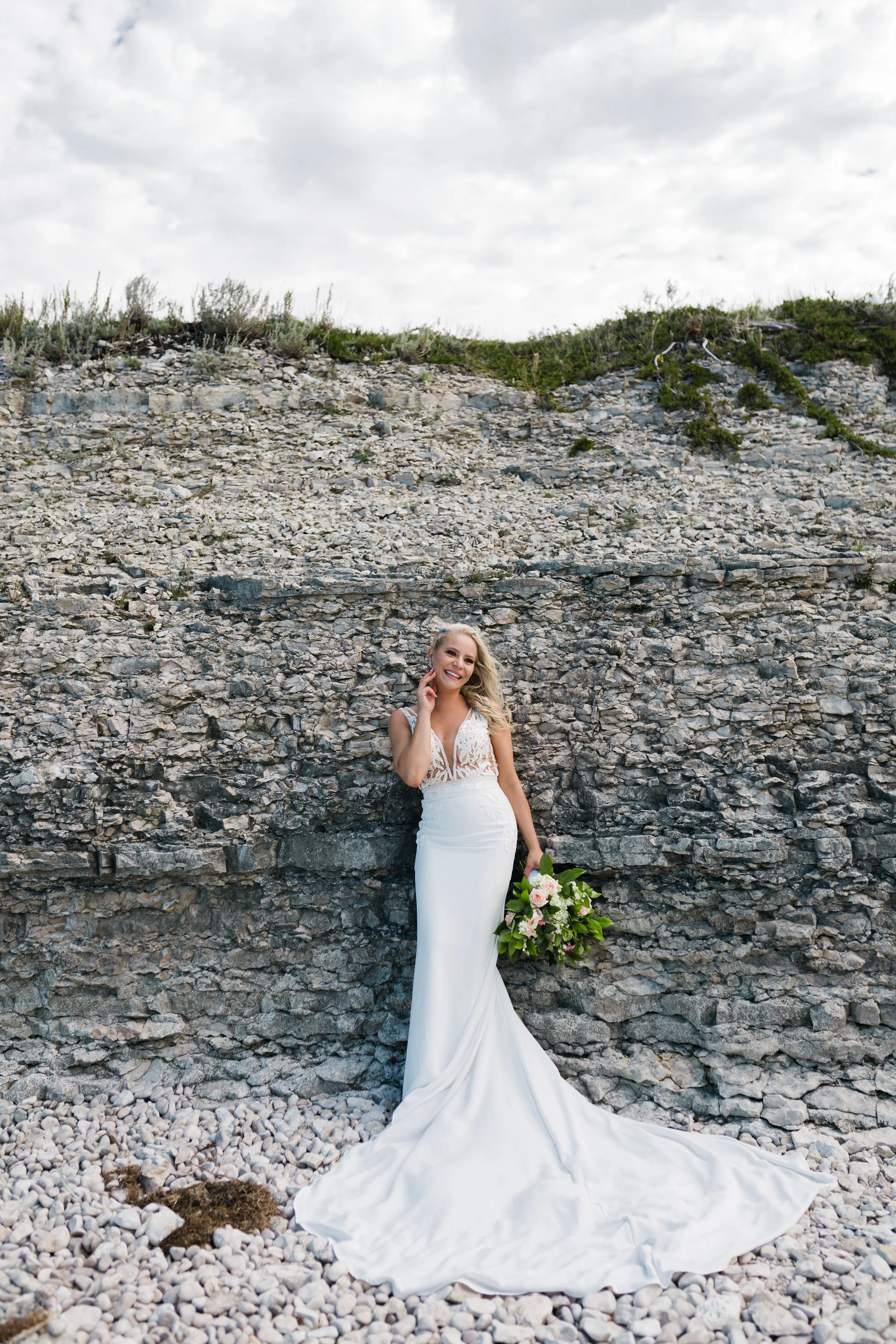 A smiling bride in a white wedding gown holding a bouquet of pink and white flowers, standing on a rocky beach with a stone wall and cloudy sky background.