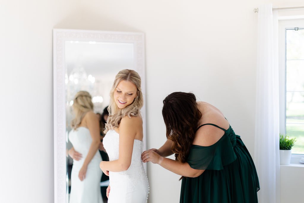 A woman in a white dress is being helped with her dress by another woman in a dark green dress inside a bright room with a mirror and window.
