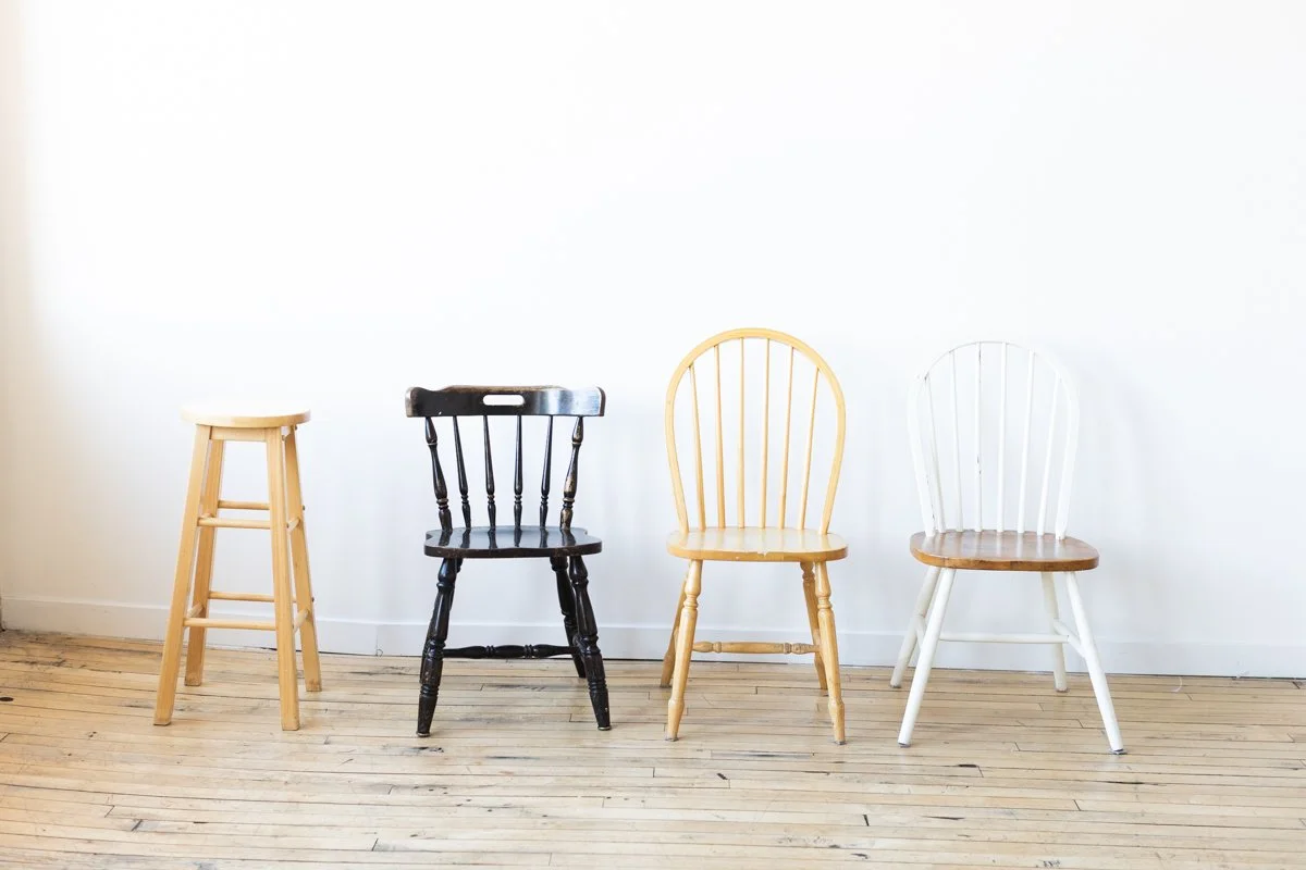 Four different chairs and a stool placed in a row against a plain white wall with a wooden floor.