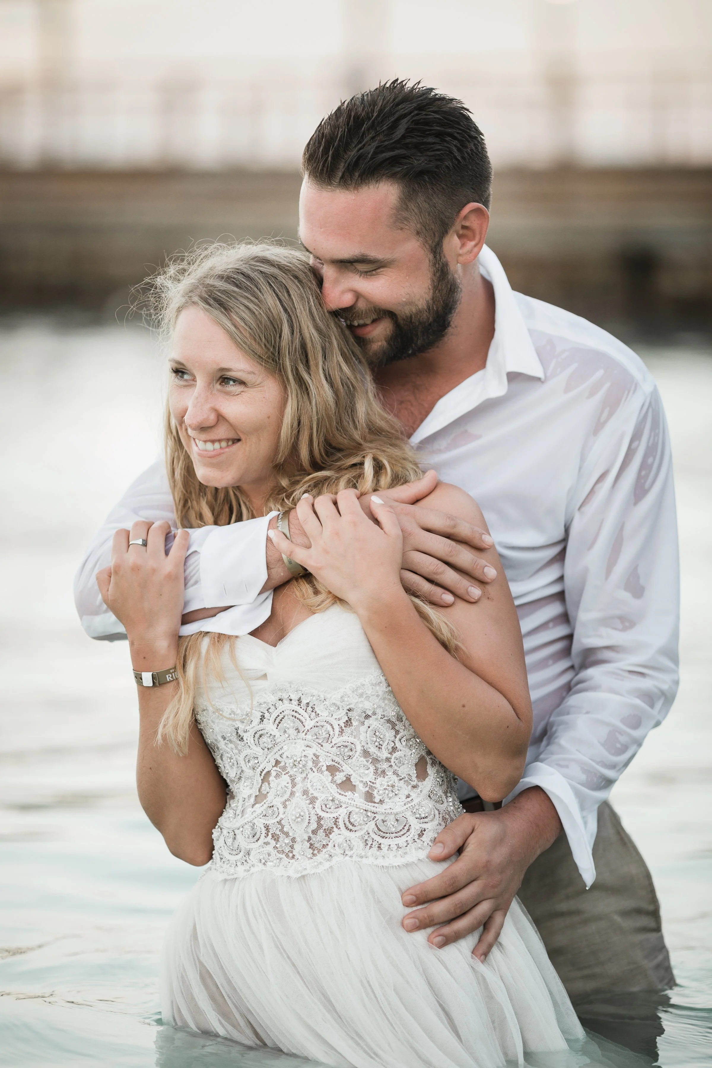 A man and woman embrace in shallow water, smiling warmly. The woman wears a white lace dress, and the man wears a white shirt. They are happy and affectionate.