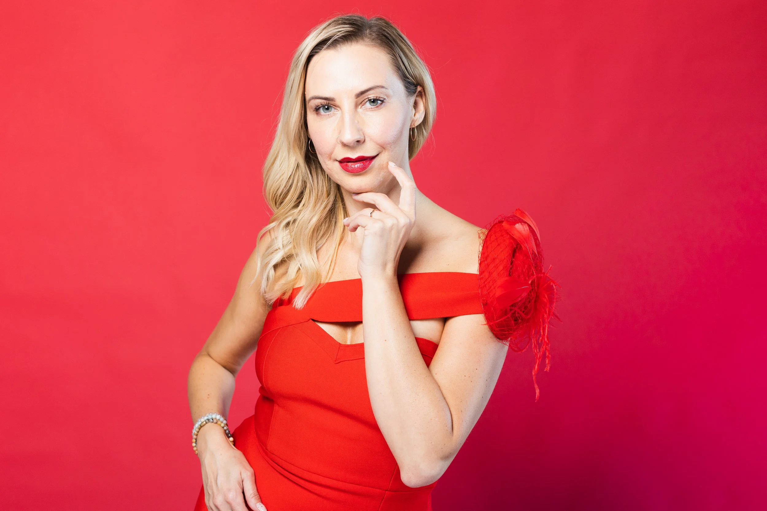 A woman with blonde hair in a red dress with an off-the-shoulder design and decorative sleeve stands against a red background. She has one hand near her chin and is looking at the camera.