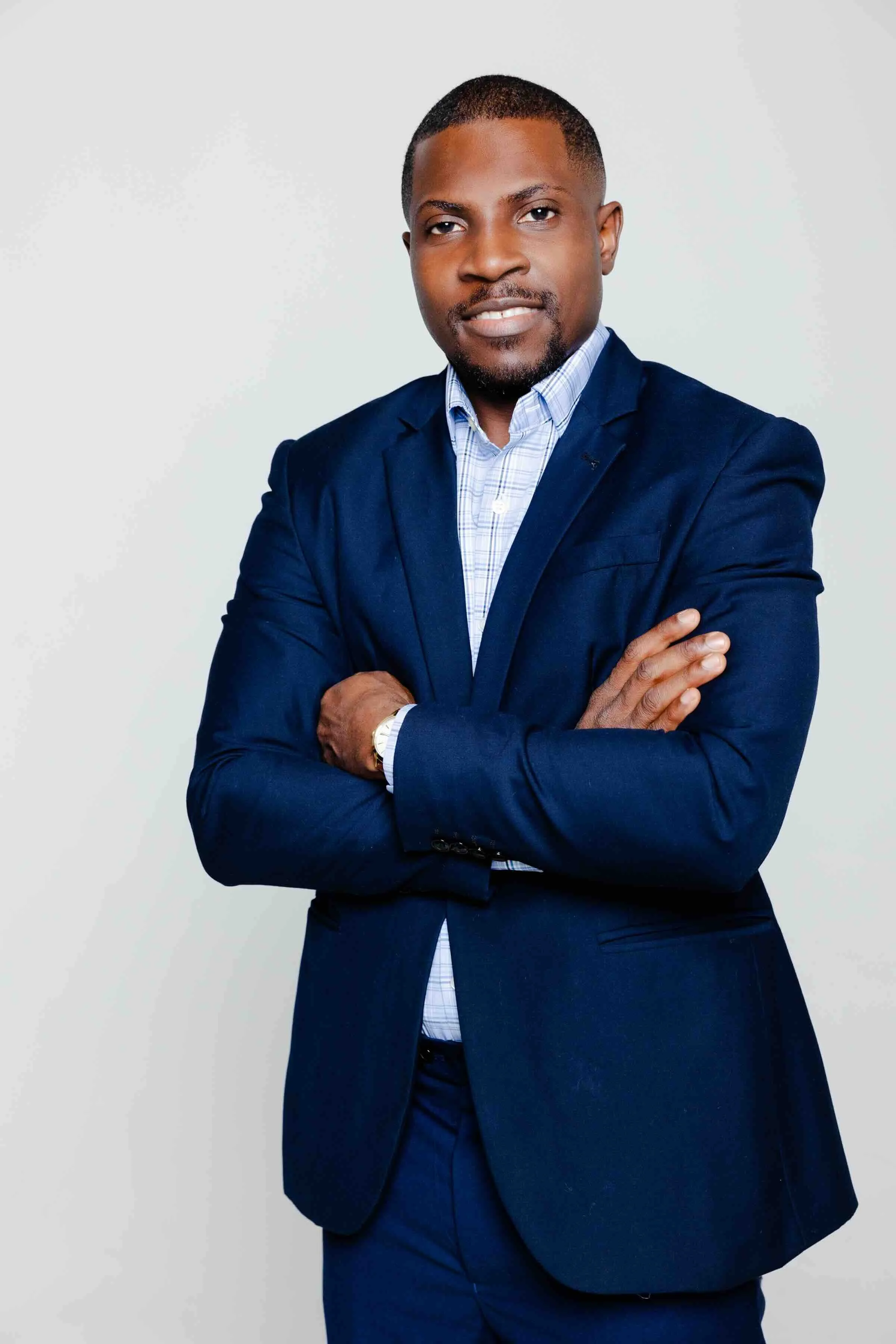 A man in a dark blue suit with crossed arms, wearing a light blue checkered shirt, standing against a plain gray background.