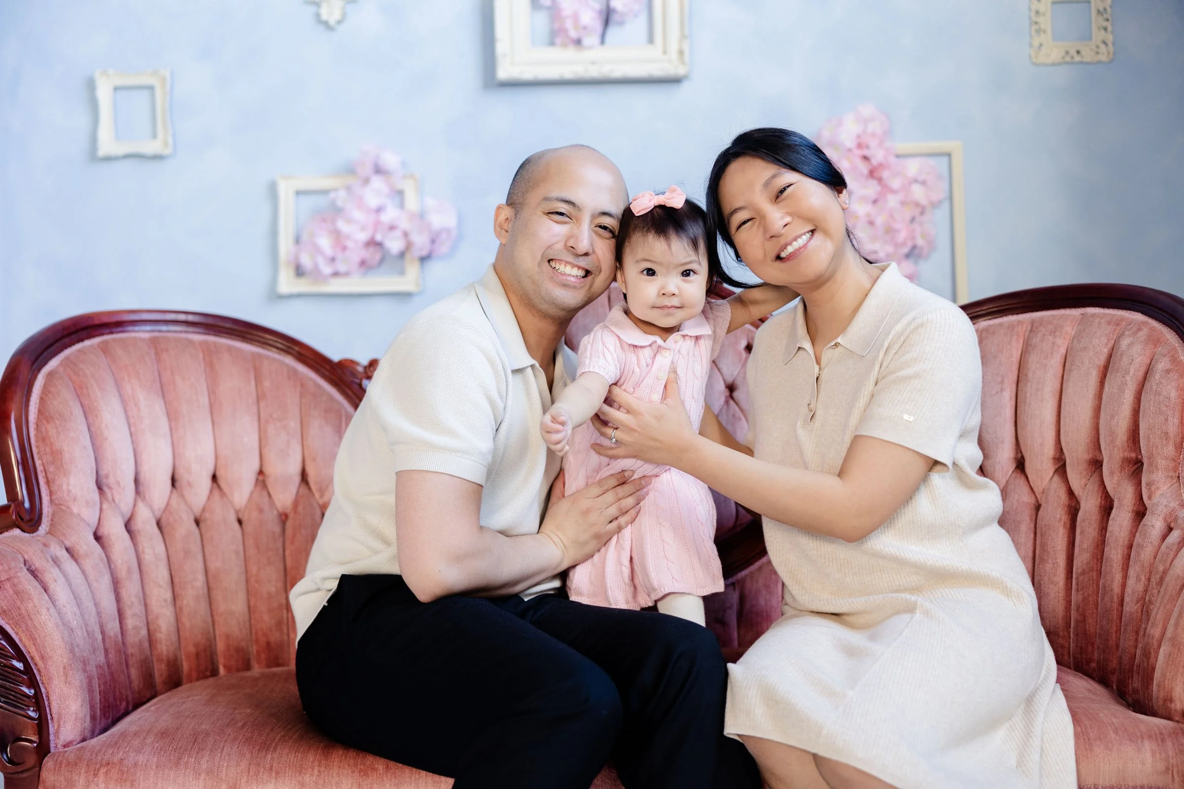 A living room with a gray tufted sofa adorned with pastel-colored pillows. A side table holding a plant stands on the left. The background features white paneled walls with a round wicker mirror, a small astronomical telescope, a table lamp, and a decorative vase with dried flowers. A woven beige rug lies on the floor in front of the sofa.