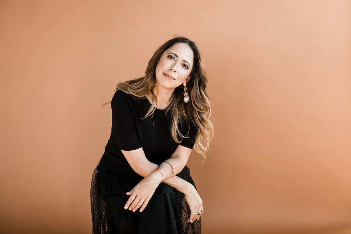 A woman with long wavy hair wearing a black top and a black skirt with a subtle pattern, sitting against a plain beige background, looking at the camera with a slight smile.