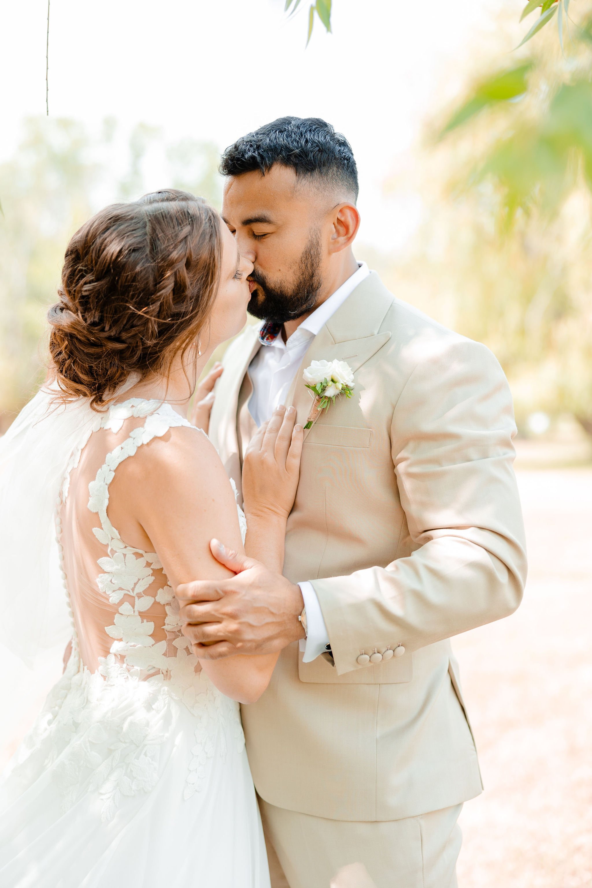 A bride and groom share a kiss outdoors on their wedding day, with the bride touching the groom's chest and the groom holding the bride's arm, amidst a background of trees.