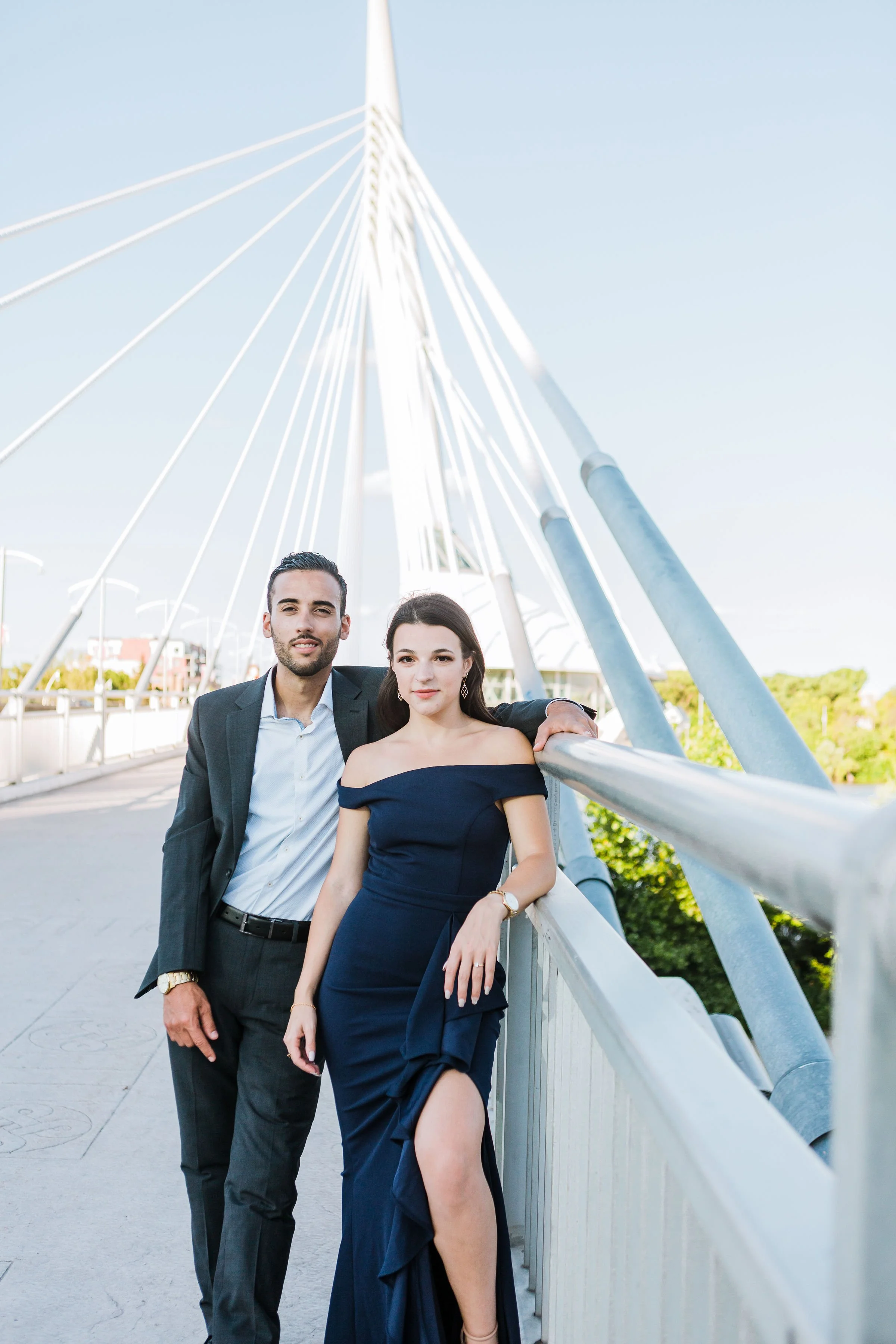 A man in a suit and a woman in a navy blue dress standing on a bridge with a modern white cable-stayed support structure, outdoors on a clear day.