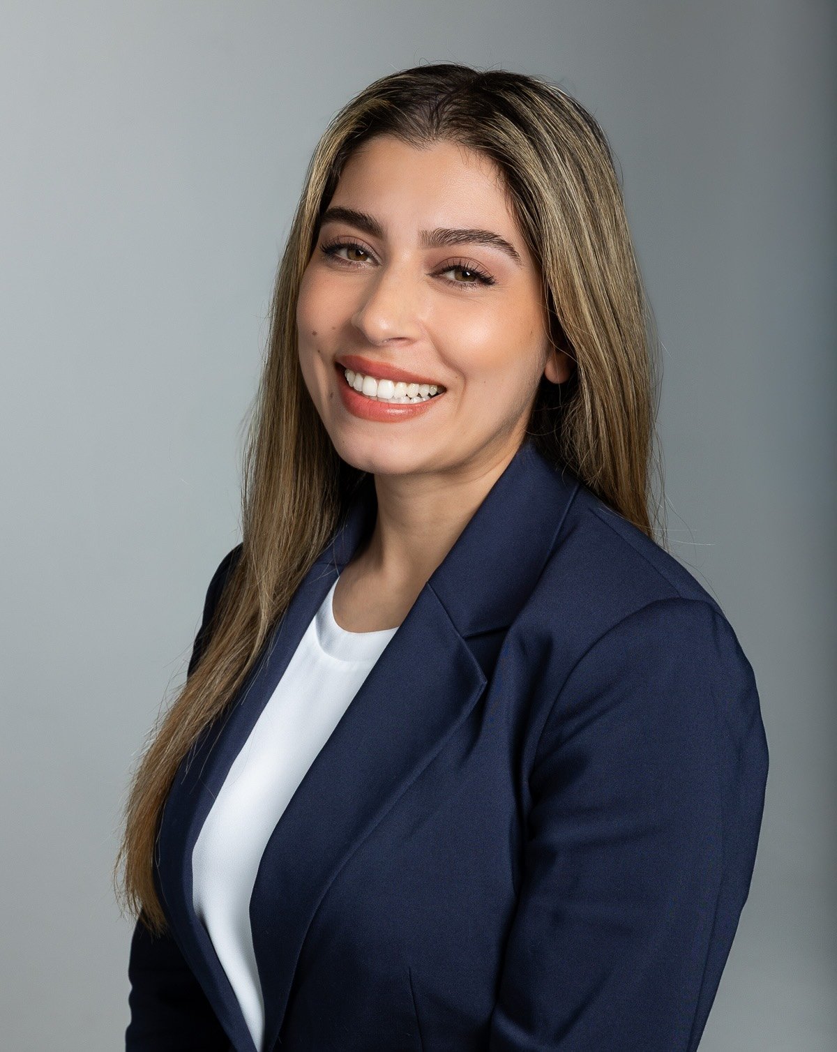A woman with long brown hair wearing a navy blazer and white top, smiling against a plain gray background.