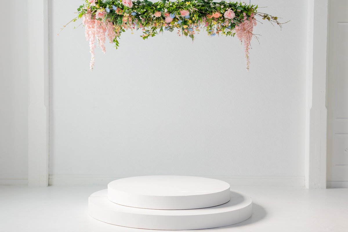 Minimalist living room with white walls and flooring, featuring a blue velvet armchair with a white blanket, a round marble and glass coffee table on a tan flower-shaped rug, a green velvet chair with pink flowering plants in white pots, a fireplace mantel, and a large green plant in a woven basket.