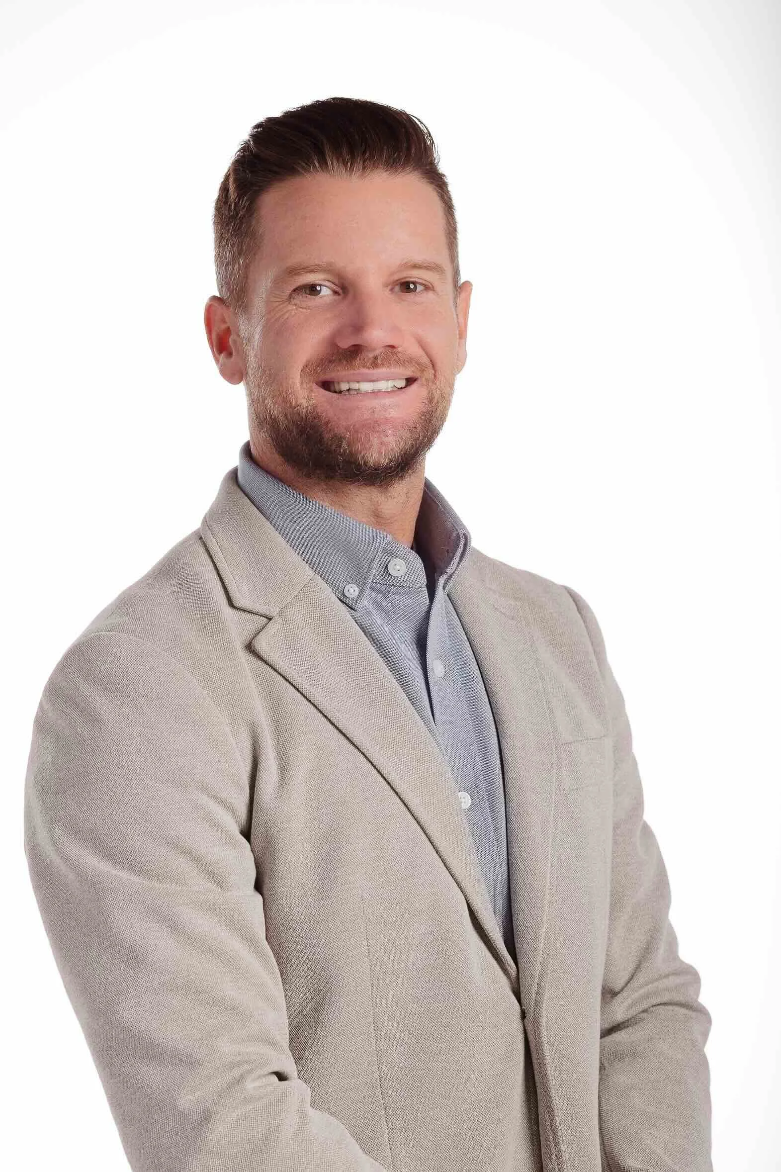 A man with light skin, brown hair, and a beard, smiling, wearing a beige blazer over a gray collared shirt, posing against a plain white background.