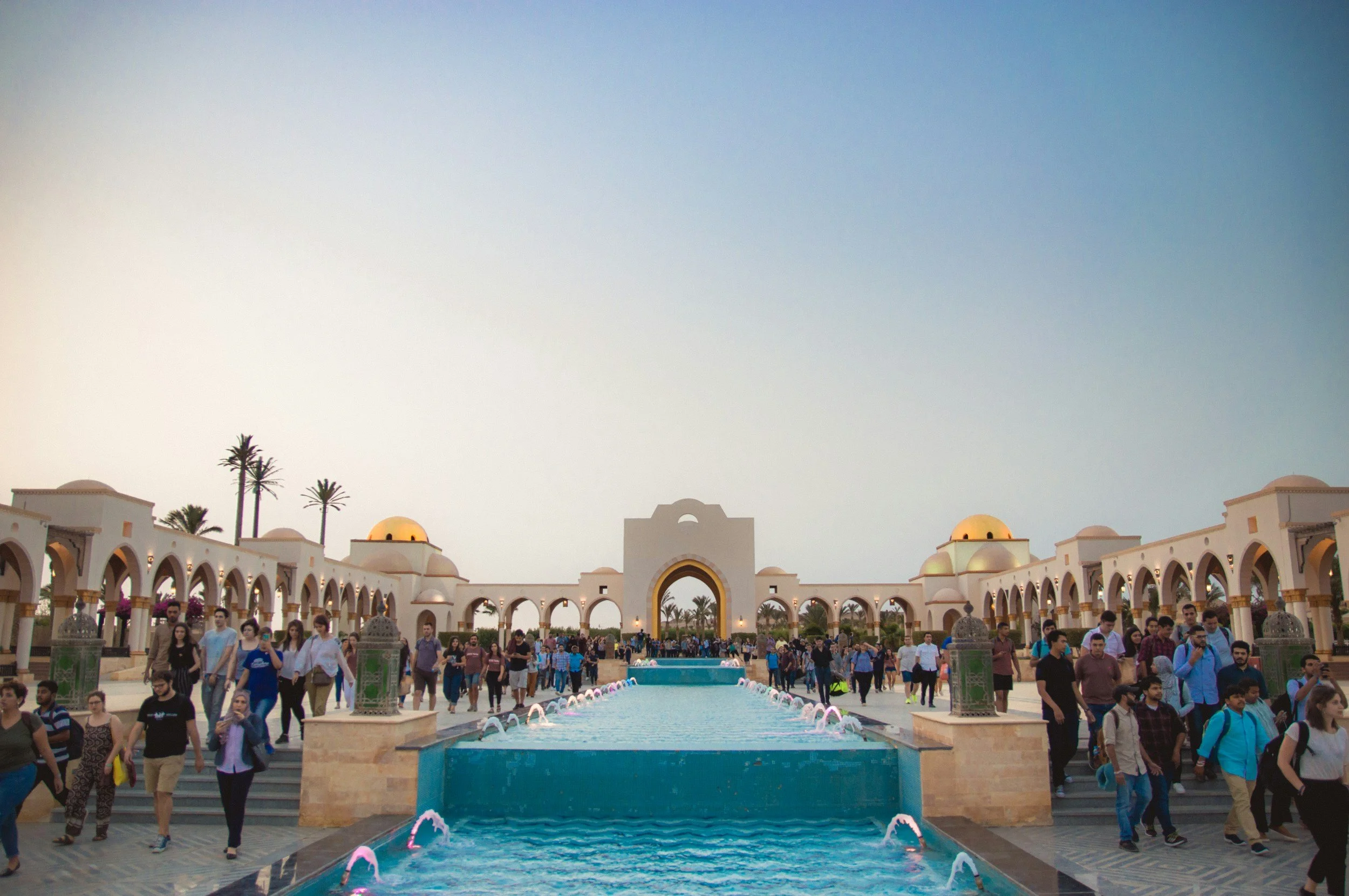 People walking around a large, open courtyard with a central fountain, surrounded by white arches and domed structures, with palm trees in the background, during dusk.