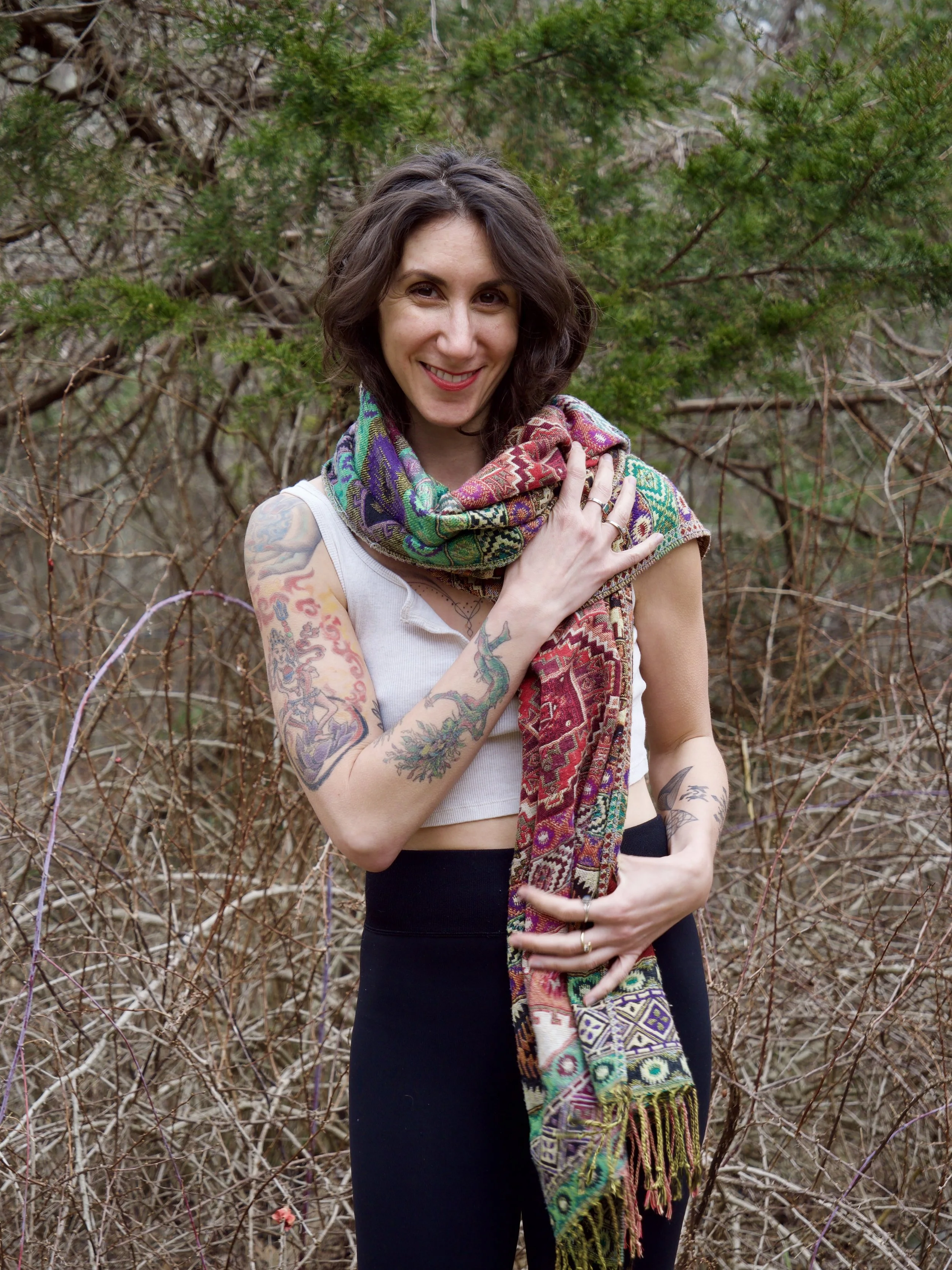 Ronni Clara Rose smiling with tattoos on her arms, wearing black pants, a white crop top, and a colorful patterned scarf around her neck, standing outdoors in front of dry bushes and green trees.