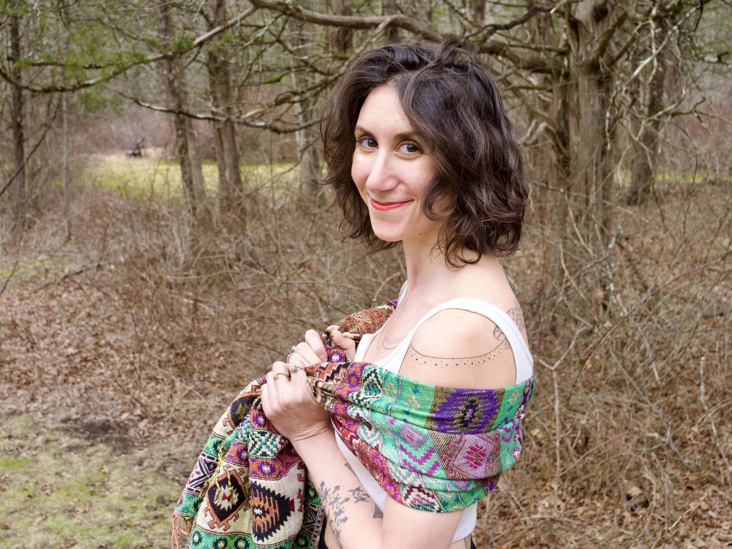 Ronni Clara Rose, a woman with dark, curly hair smiling outdoors in a wooded area, holding a colorful patterned scarf.
