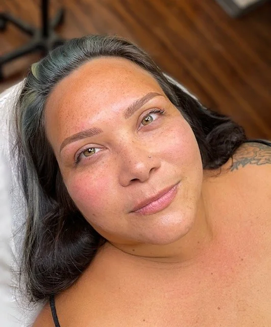 Close-up of a woman lying down with her head tilted slightly, showing her face and shoulders. She has dark hair, light skin with freckles, and a slight smile.