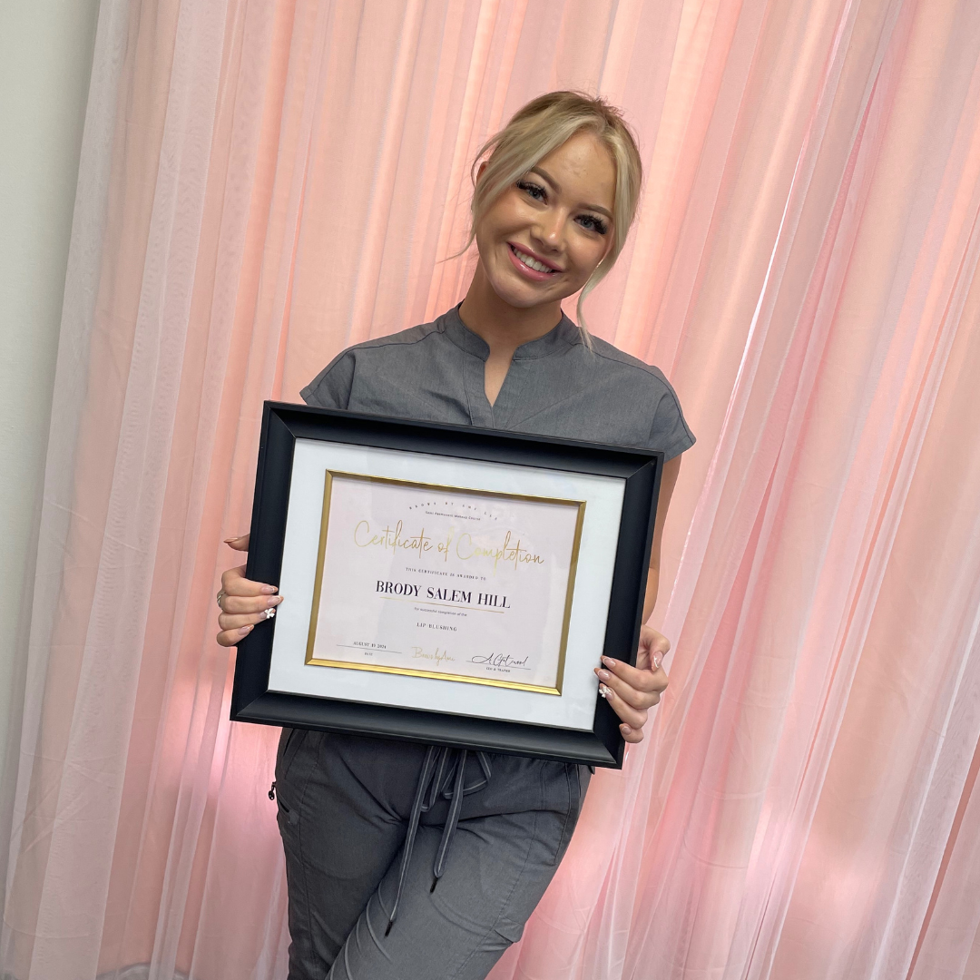 A woman with blonde hair smiling and holding a framed certificate of completion, standing in front of pink curtains.