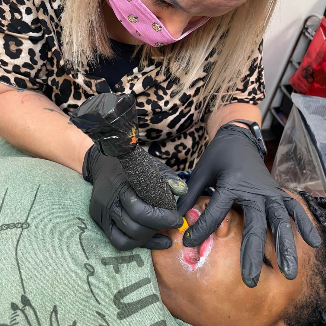 Tattoo artist wearing a leopard print shirt and a pink face mask with cookie designs inkingtattoo on a person's neck using a tattoo machine. The person receiving the tattoo has dark skin, braided hair, and is lying down.