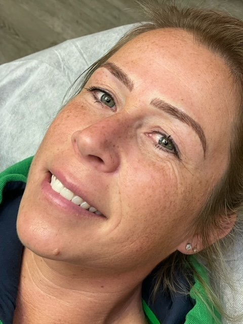 Close-up of a smiling woman with light skin, freckles, blue eyes, and brown hair, lying on a medical examination table.