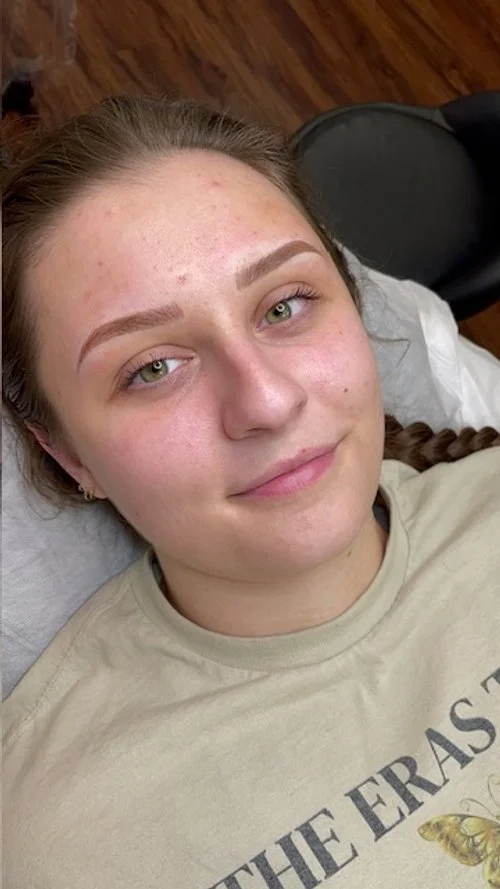 Young woman with clear skin and braided hair, lying on a bed or chair, smiling slightly, with a wooden floor and black chair in the background.