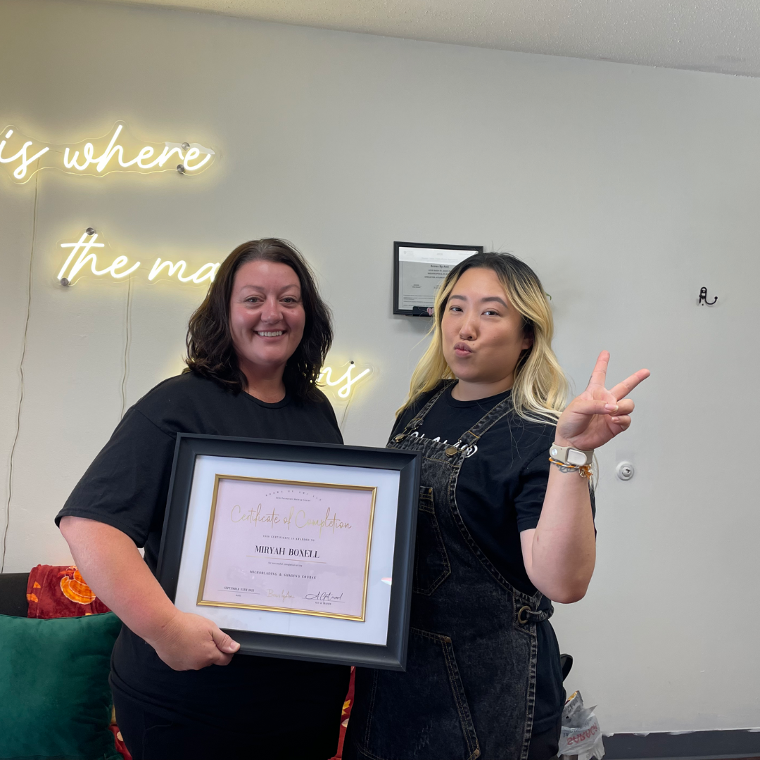 Two women smiling, one holding a framed certificate, the other making a peace sign with her fingers, standing in front of a wall with neon signs and a framed document.