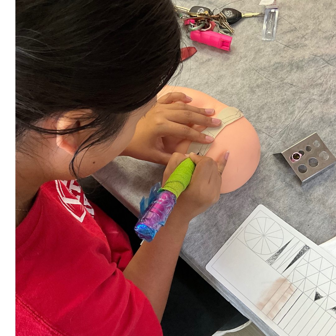 A person with dark hair in a red shirt is practicing tattooing on a silicone practice skin, using a tattoo machine with a green grip. On the table, there are keys, a pink tool, a small ruler, and a sheet of practice tattoo outlines.
