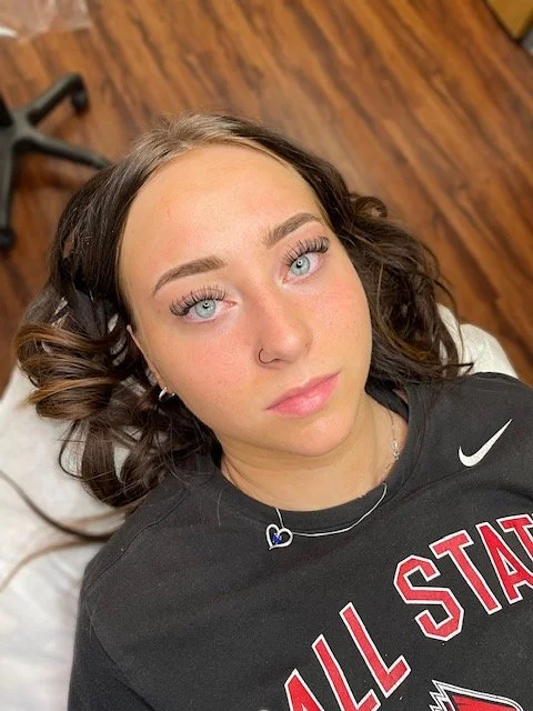 A young woman with light skin and blue eyes lying on a white surface, wearing a black All Star t-shirt with a Nike logo, a necklace with a heart-shaped pendant, and a nose ring.